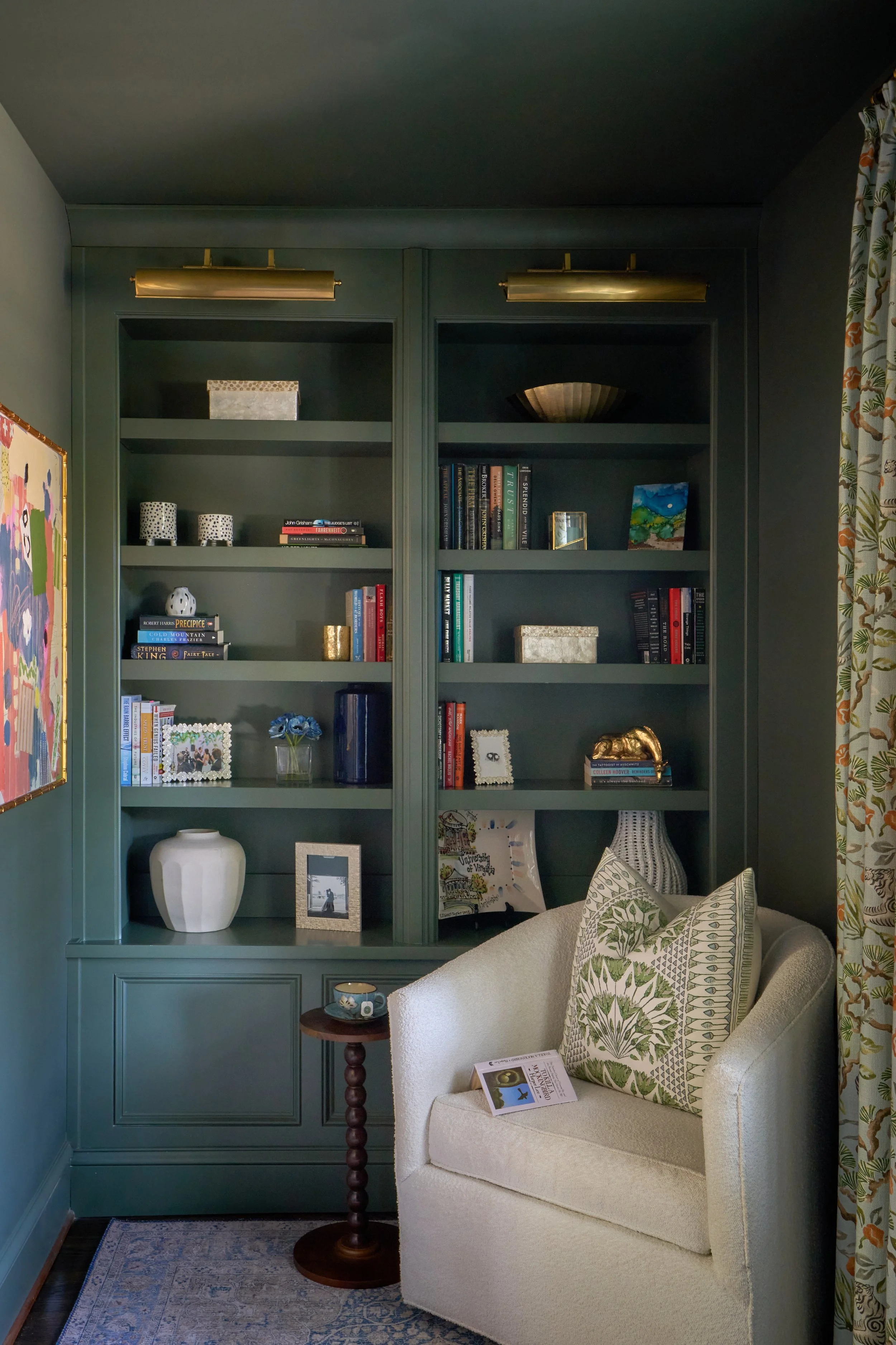 A cozy reading nook with a green built-in bookshelf filled with books, decorative items, and framed photos. An off white armchair with a patterned pillow is positioned beside a small wooden cocktail table, and a decorative rug on the dark wood floor.
