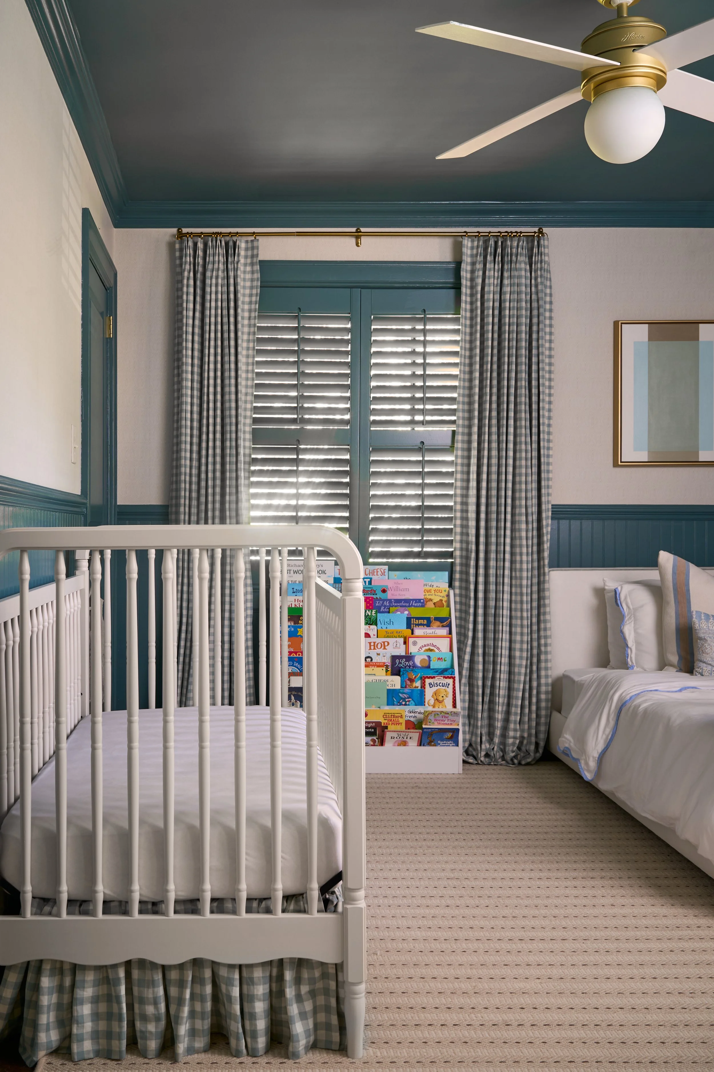 A white cribs with a bed, adjacent to a white hospital-style crib, a bookrack filled with children's books, a window with blue shutters, and blue and white checkered curtains, in a children's bedroom, with beige carpet and framed artwork on the wall,
