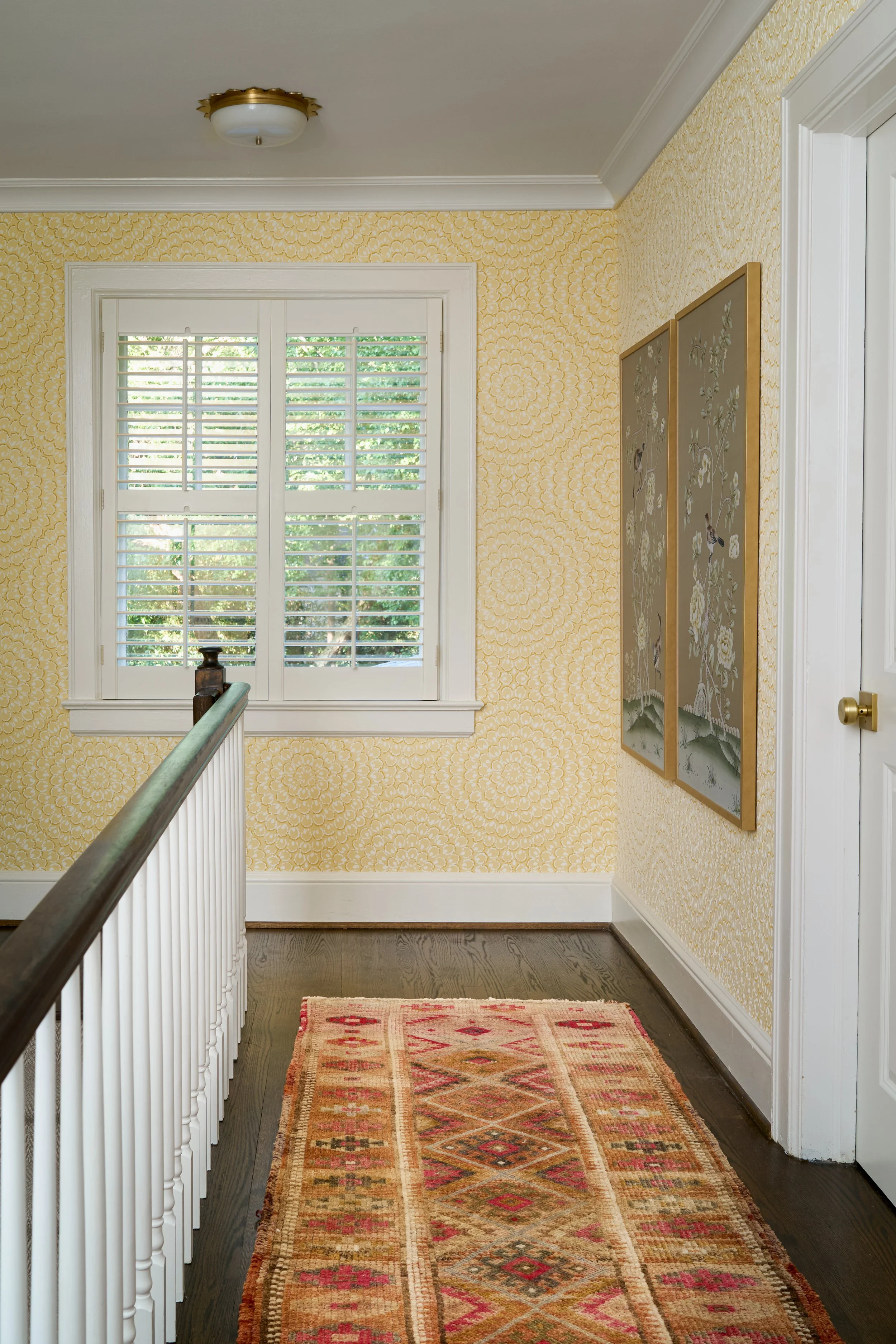 Yellow thibaut wallpaper is carried from the foyer to the upstairs hallway. Beautiful sun shaped semi-flushmount, two chinoiserie inspired hand painted panels are hung on the wall and a pink, yellow, white and cream runner complete the space.