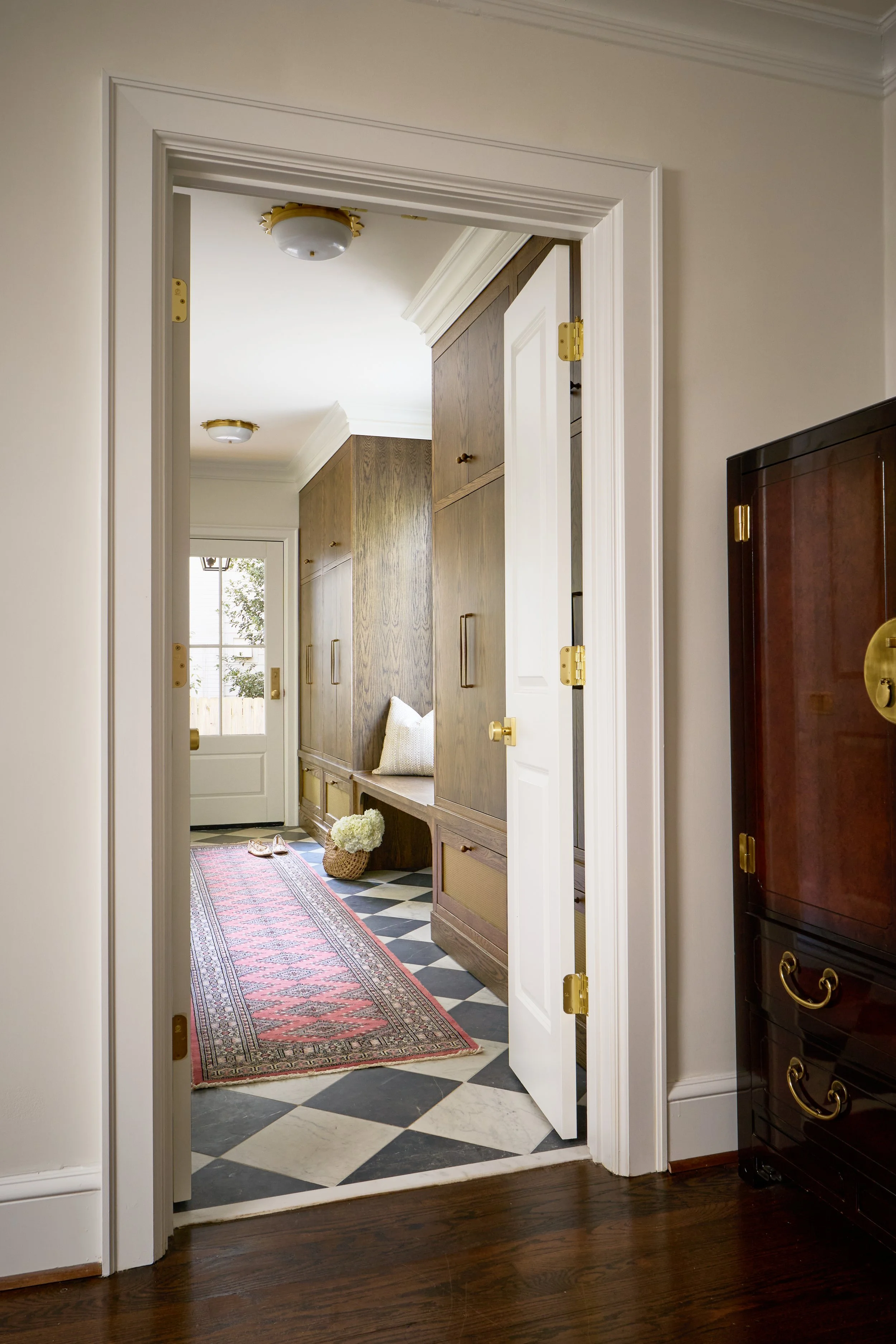 View through a doorway into a hallway with wooden cabinets, a patterned rug, a wicker basket with flowers, and a pair of shoes on the floor. The hallway has a black and white checkered tile floor, a window, and white walls with crown molding.