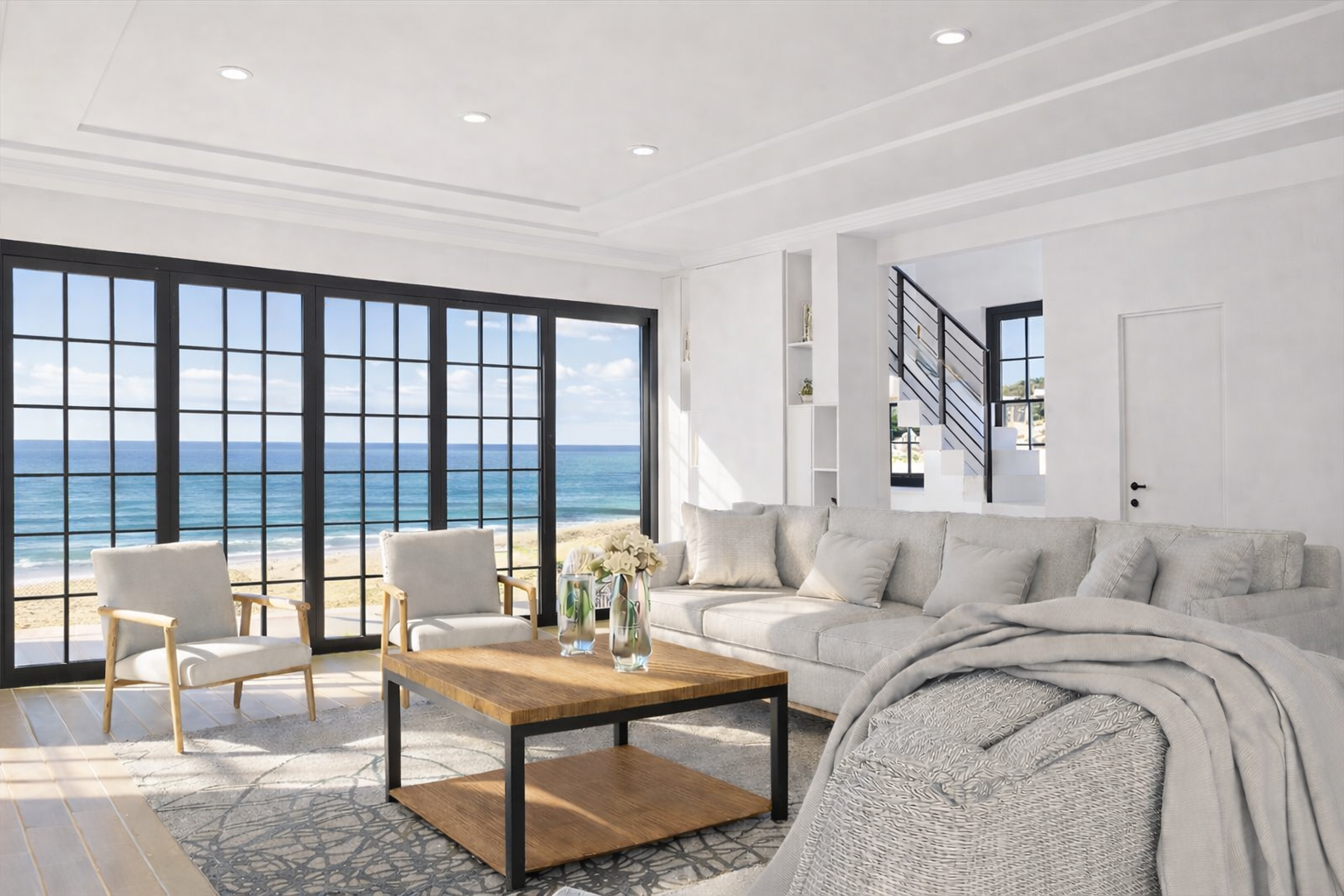 Bright living room with large windows overlooking the ocean, furnished with a sectional sofa, armchairs, a wooden coffee table with vases of flowers, and a light-colored area rug.