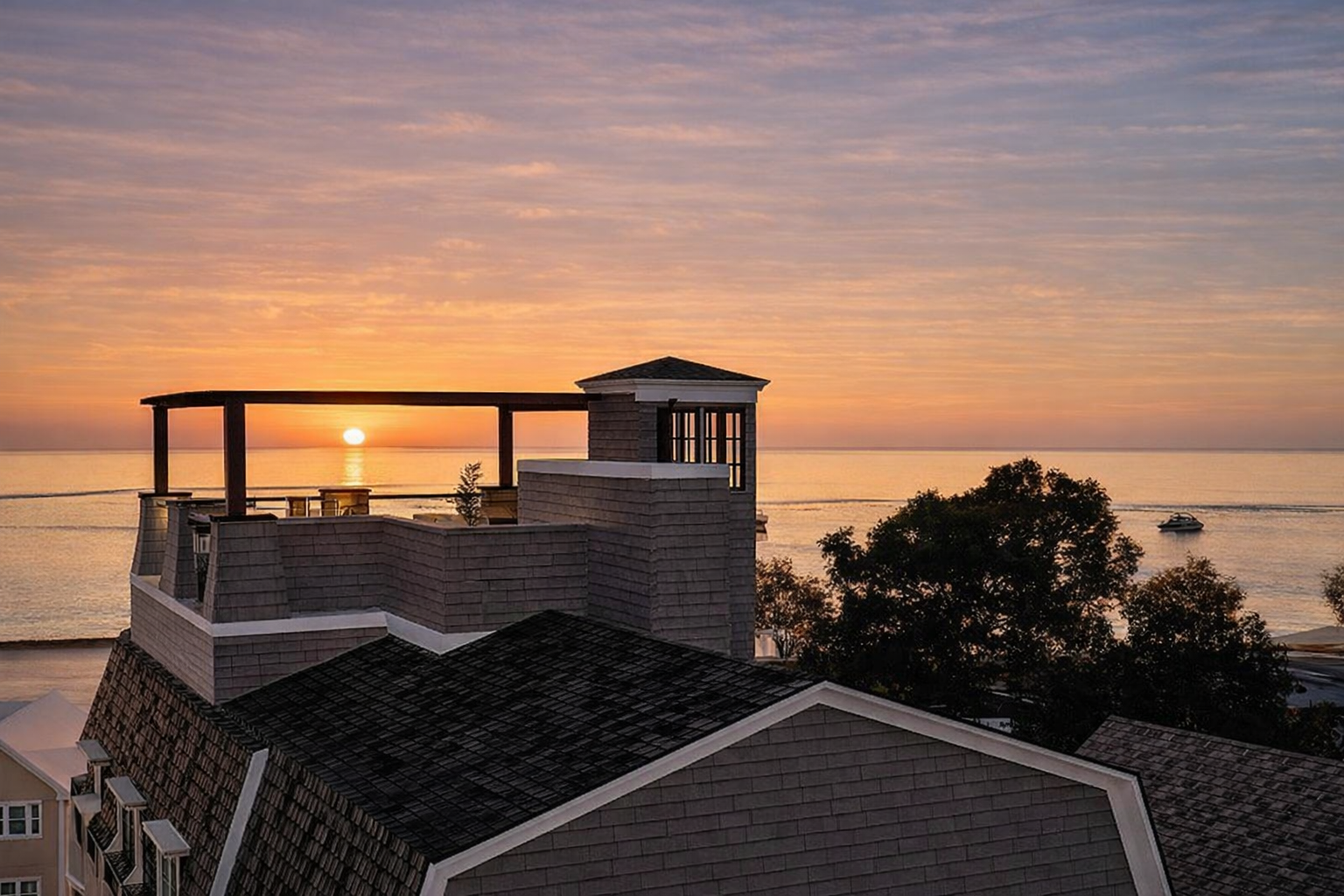 Sunset over the ocean viewed from a rooftop terrace with a small building and trees in the foreground.