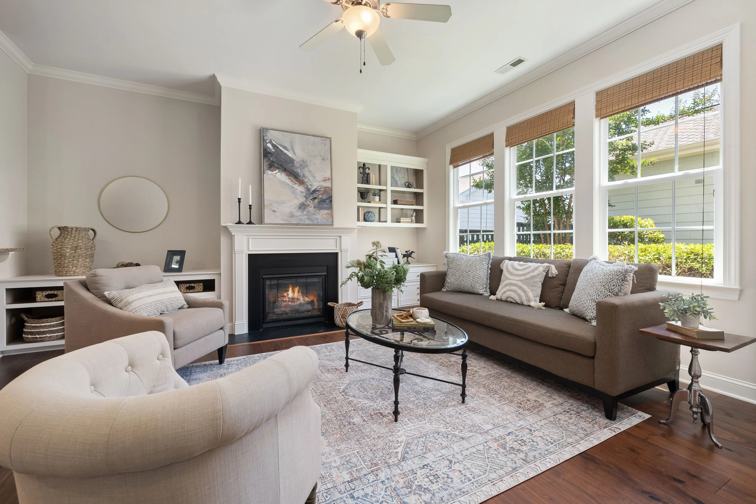 A cozy living room with a beige curved sofa, a rectangular glass coffee table, a fireplace with artwork above it, multiple windows with wooden blinds, a ceiling fan, and various decorative items including a plant, lamps, and pillows.