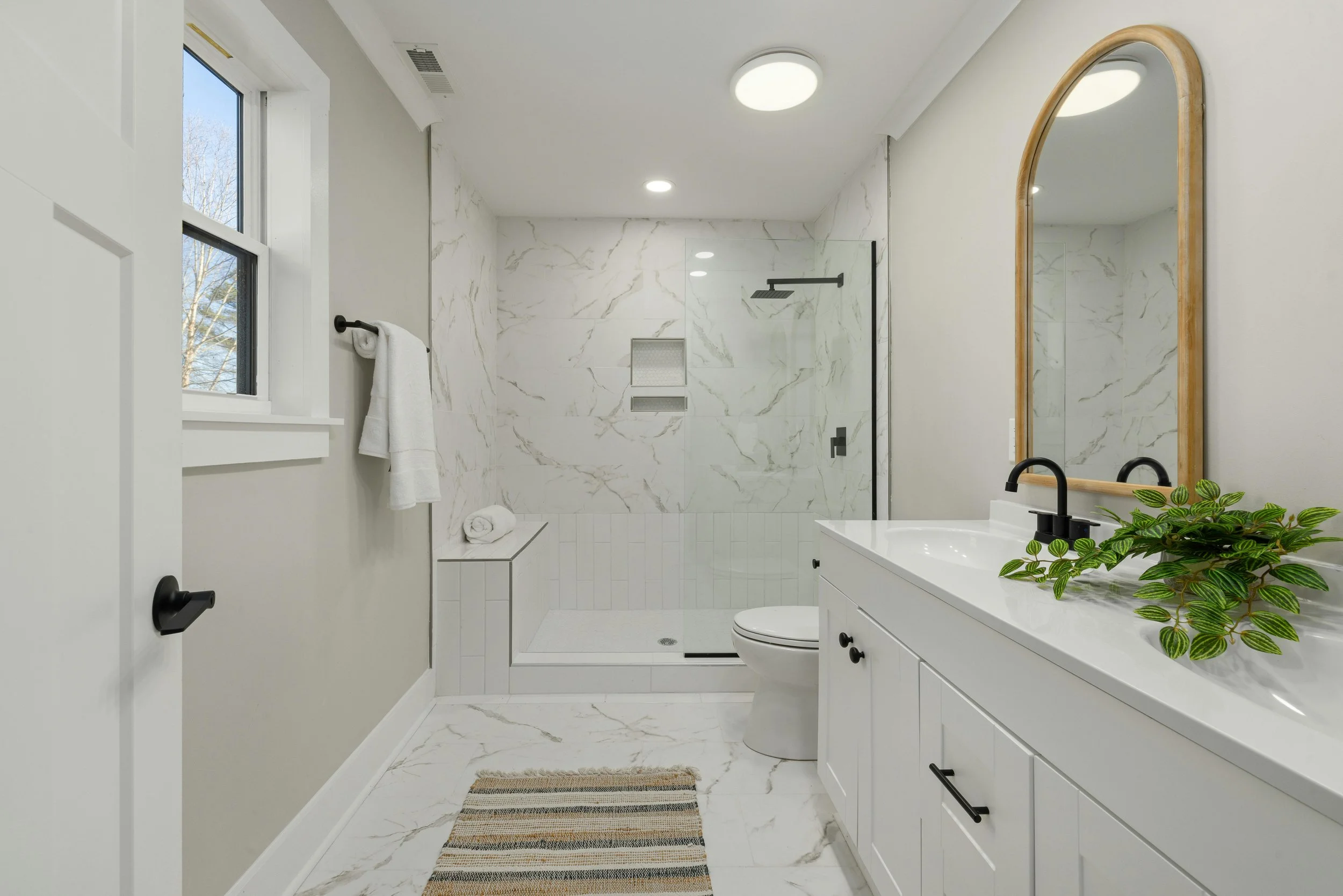 Modern bathroom with white marble walls and floors, a walk-in shower with black fixtures, a white vanity with black handles, a round mirror with a wooden frame, a striped rug, a window, and a potted plant.