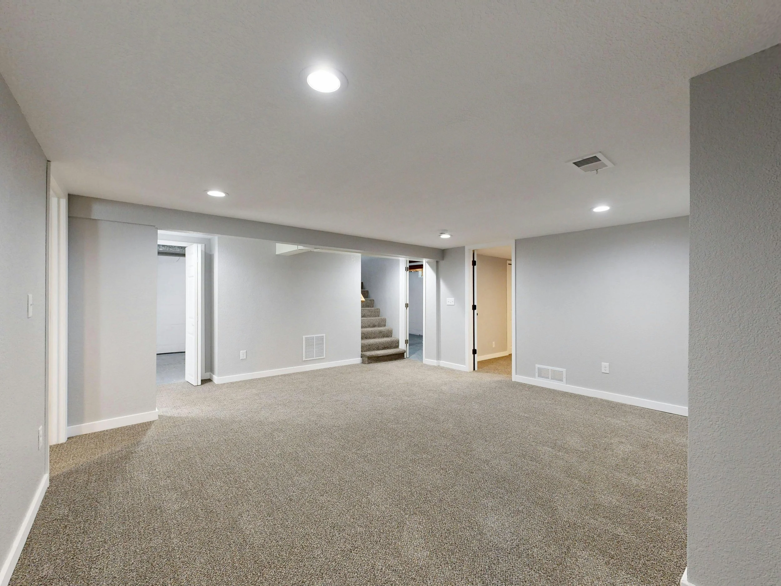 Empty living space with beige carpet, white walls, ceiling lights, stairs, and doorways.