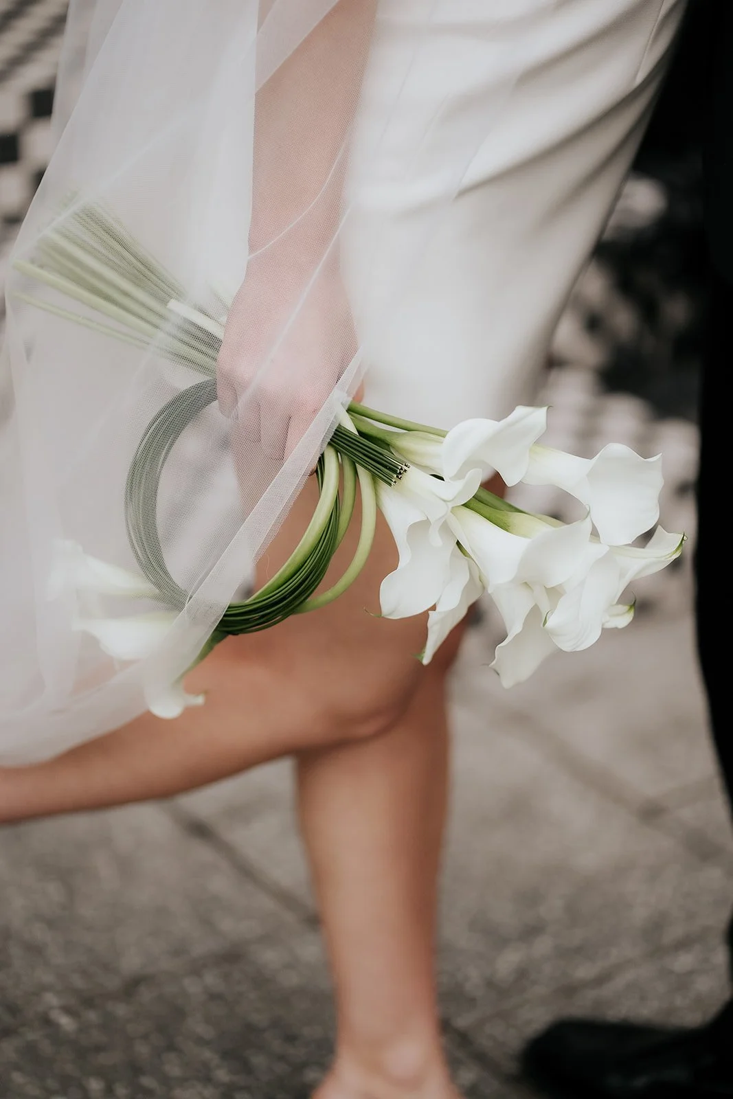 Close-up of a person holding a bouquet of white calla lilies wrapped in delicate white fabric. The focus is on the flowers and the person's hand, with a blurred background.