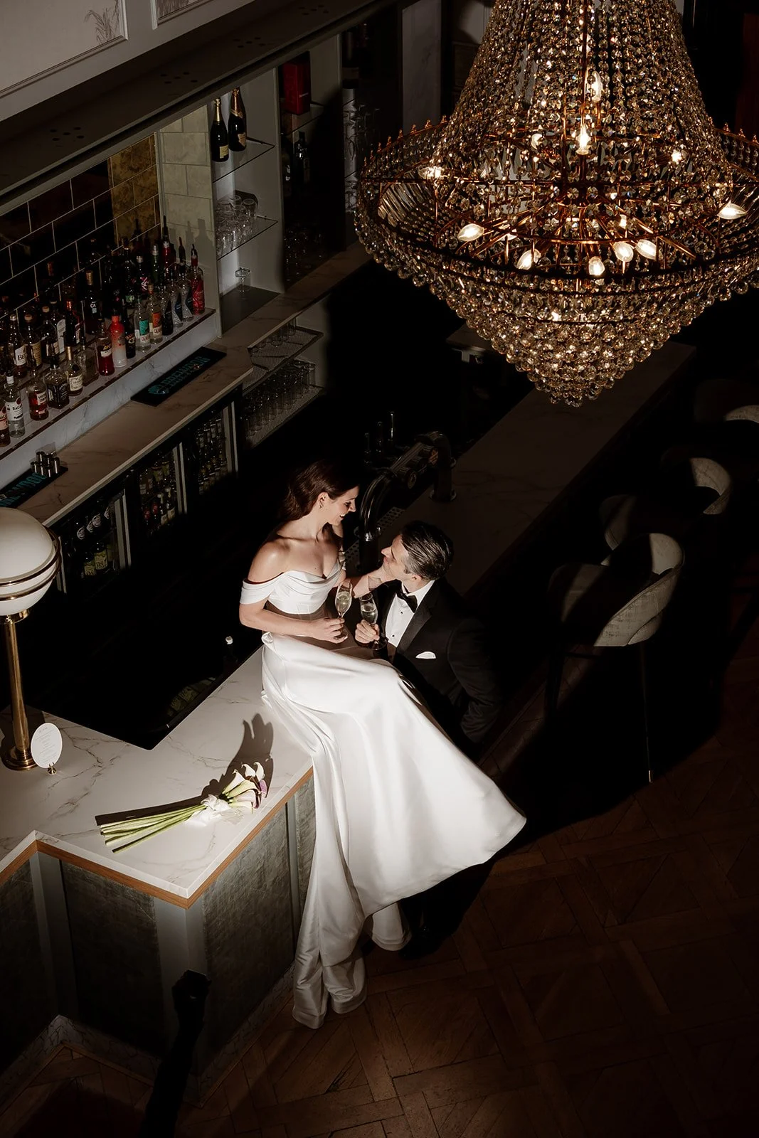A woman in a white wedding dress and a man in a tuxedo sharing a romantic moment with champagne in a luxurious bar with a large chandelier overhead.
