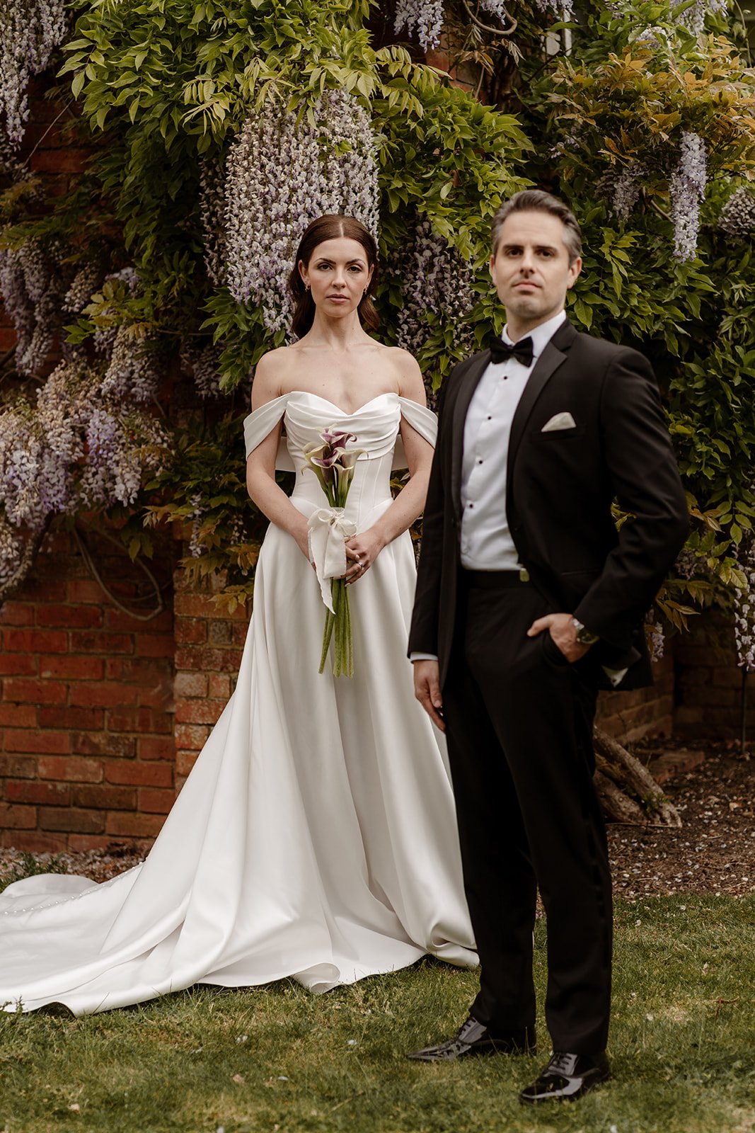A bride in a white wedding gown holding calla lilies and a groom in a black tuxedo standing outside in front of a wall of flowering wisteria and green foliage.