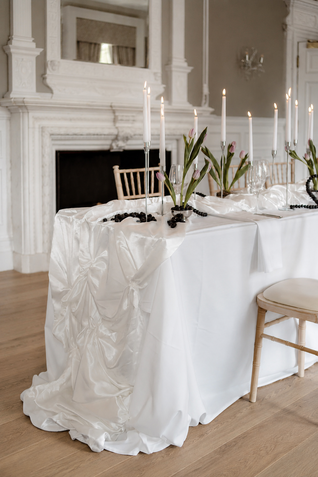 Elegant dining table decorated with white satin cloth, pink tulips in small vases, tall white candles, black beads, and empty wine glasses in a room with white ornate wall details and a fireplace.