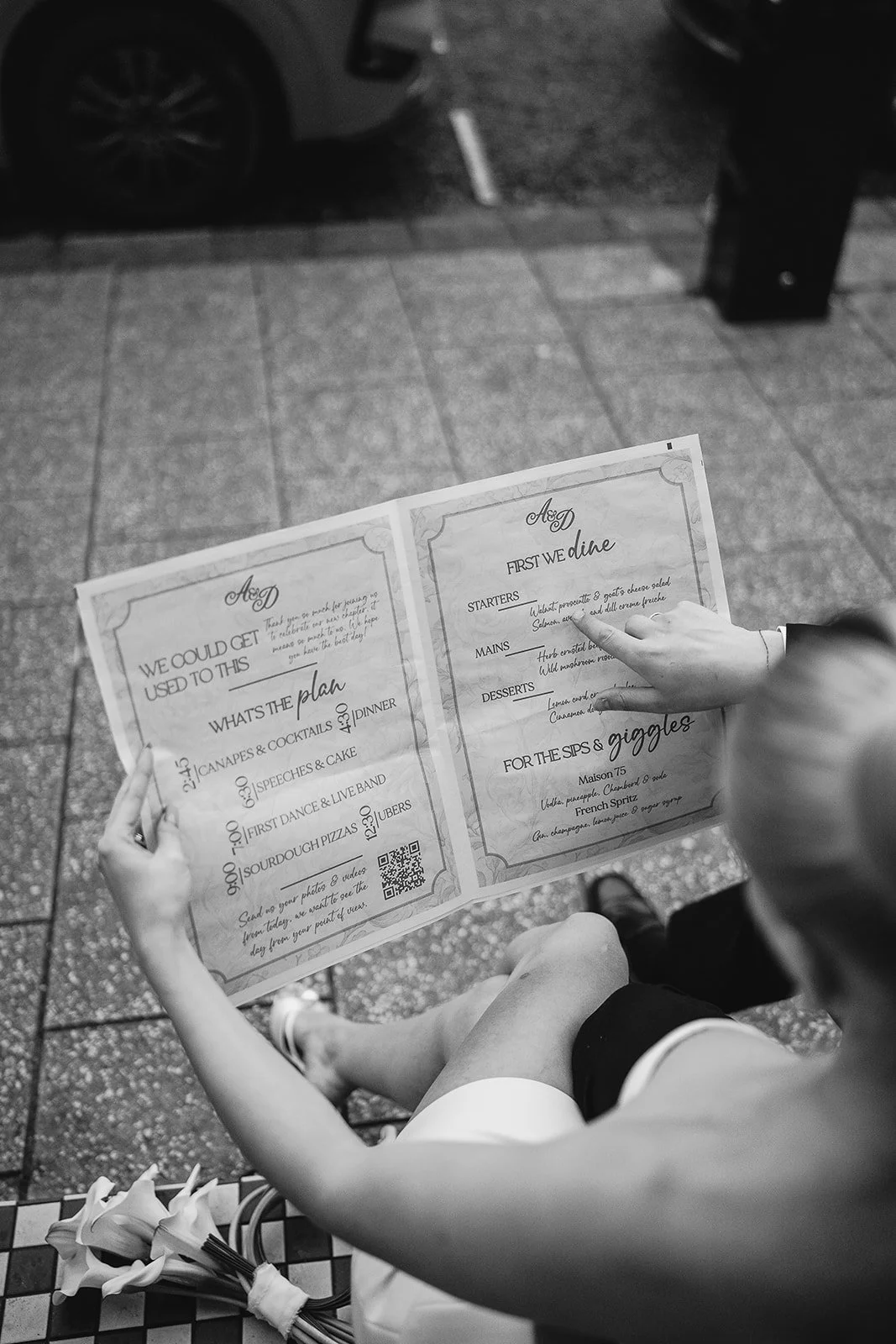 A person holding a menu outdoors on a sidewalk, with a bouquet of lilies in the foreground. The menu features a list of appetizers, main courses, desserts, and drinks in a decorative style.