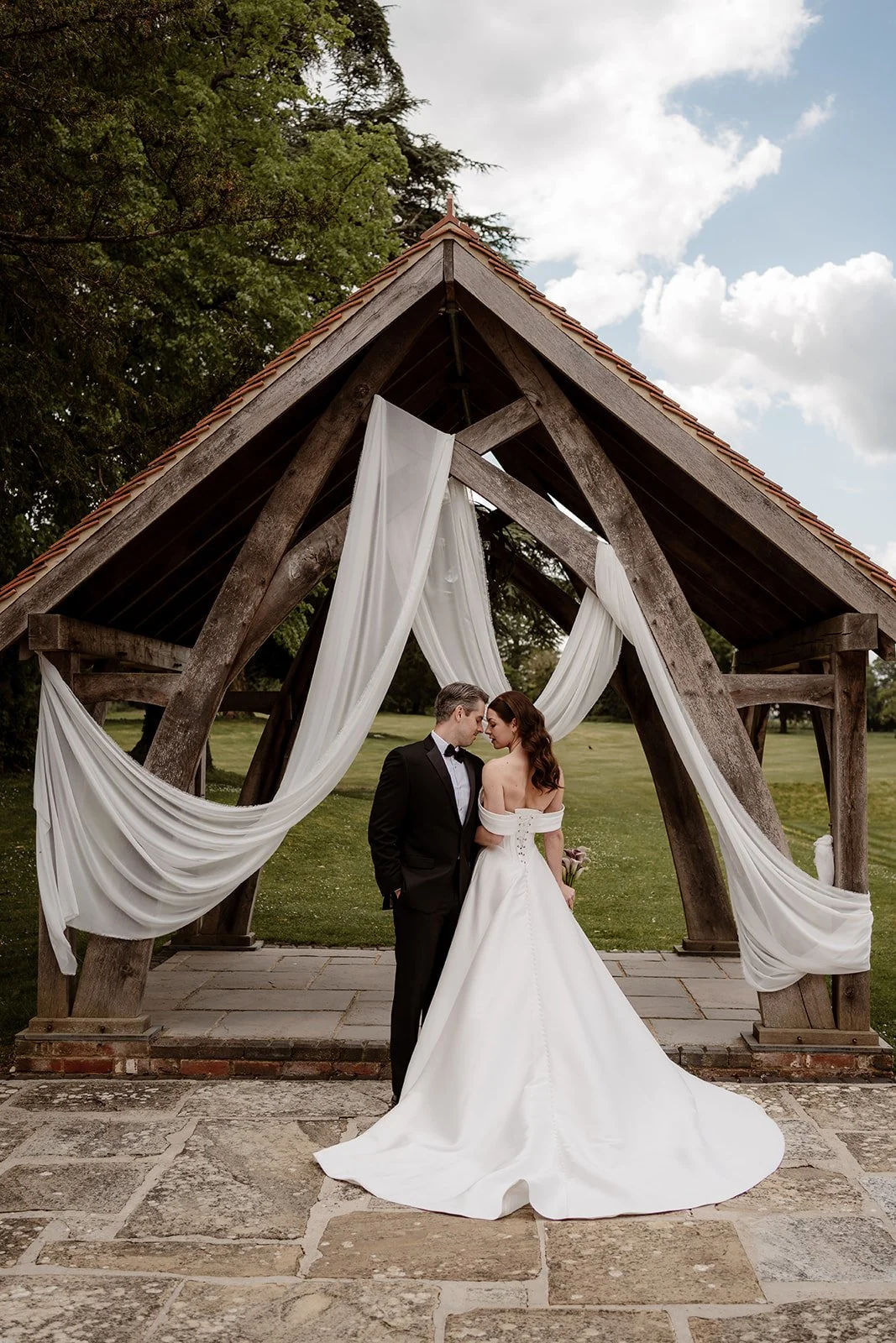 A bride and groom standing close together under a rustic wooden arch decorated with white drapes, outdoors on a cloudy day.