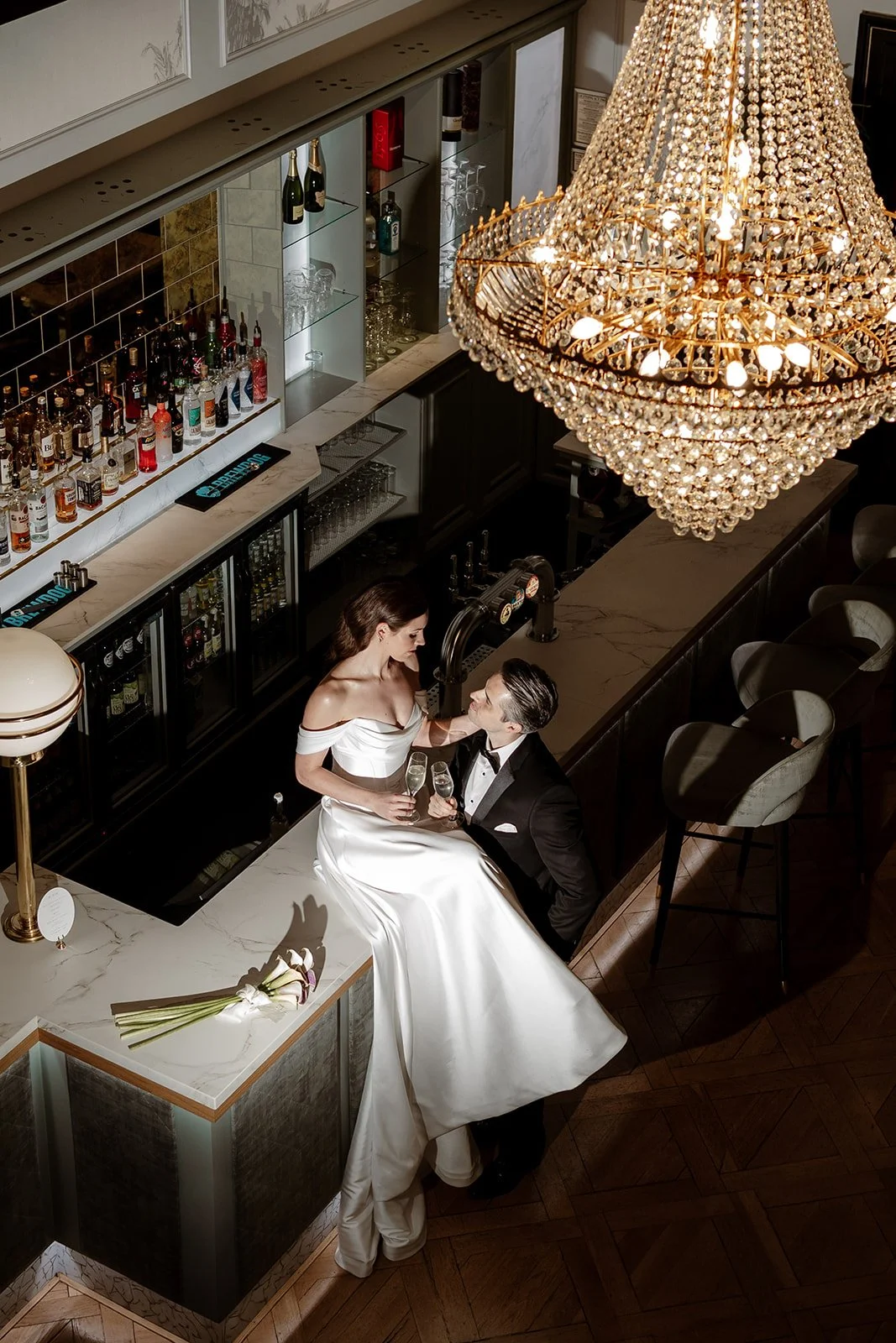 A bride and groom at a bar in a fancy venue, with a large chandelier overhead. The bride is in a white off-shoulder wedding gown, holding champagne glasses, while the groom is in a black tuxedo, sitting beside her.