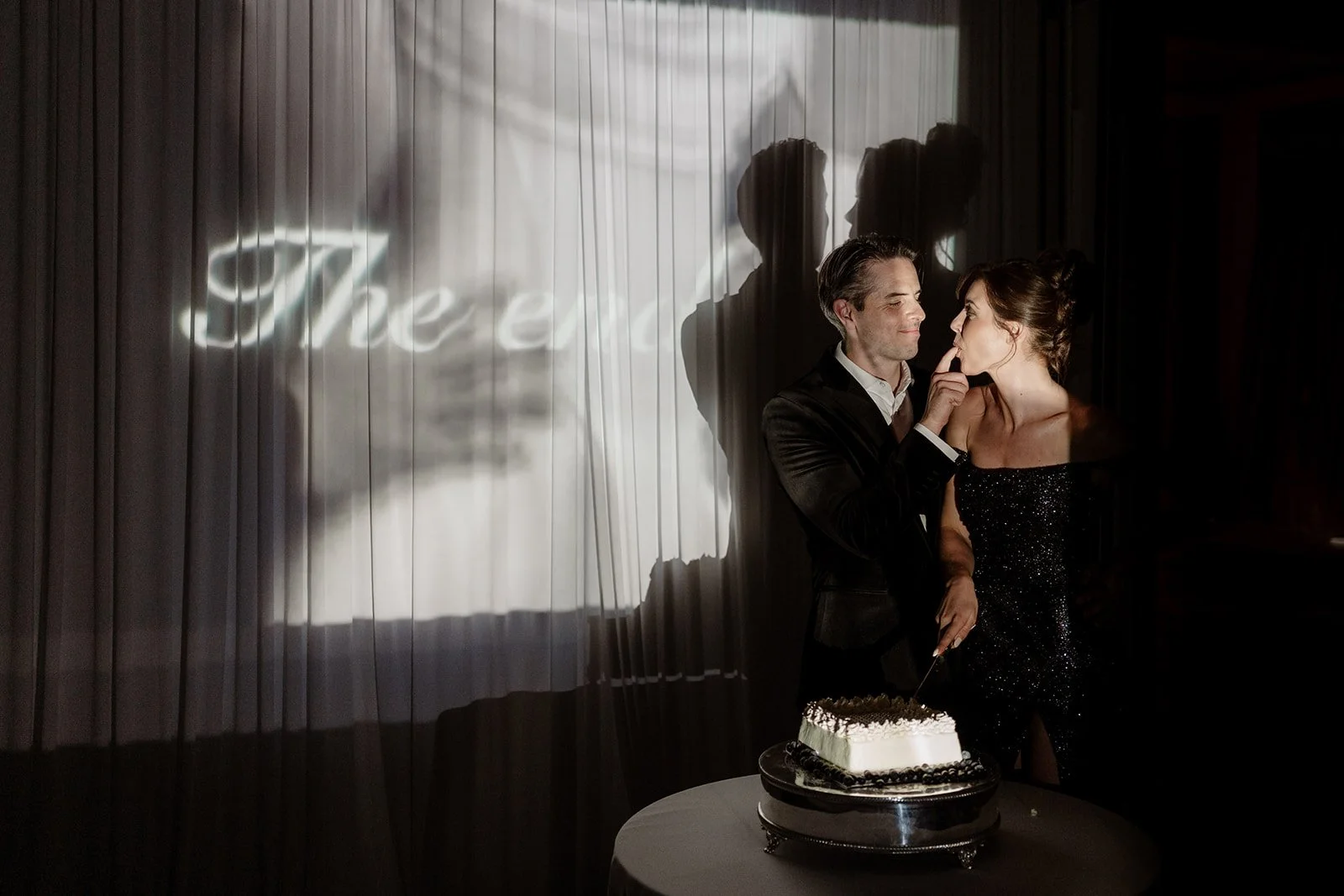 A man and woman are standing close together at a celebration, with a cake on a table in front of them. The man is gently holding the woman's face while she touches his lips with her finger. There is a shadow of a woman on a curtain behind them, with 
