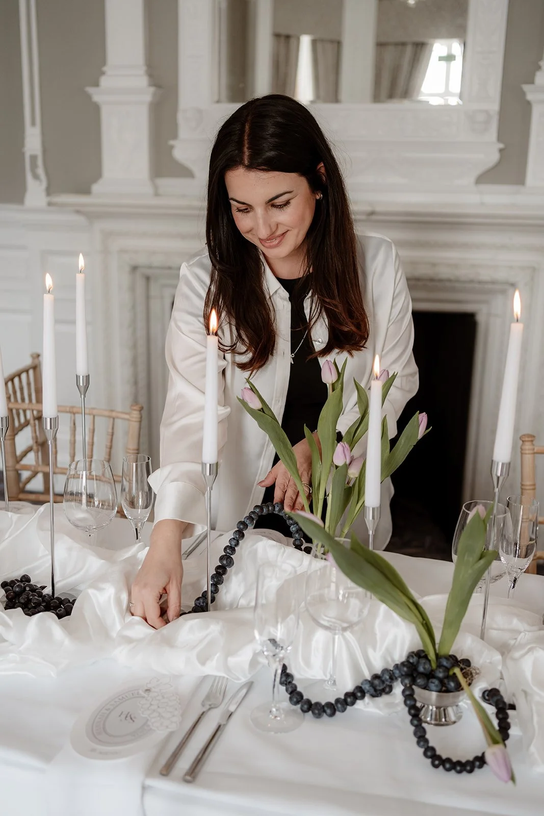 Woman setting a table with pink tulips, black beaded necklaces, and lit candles in a bright, elegant room.