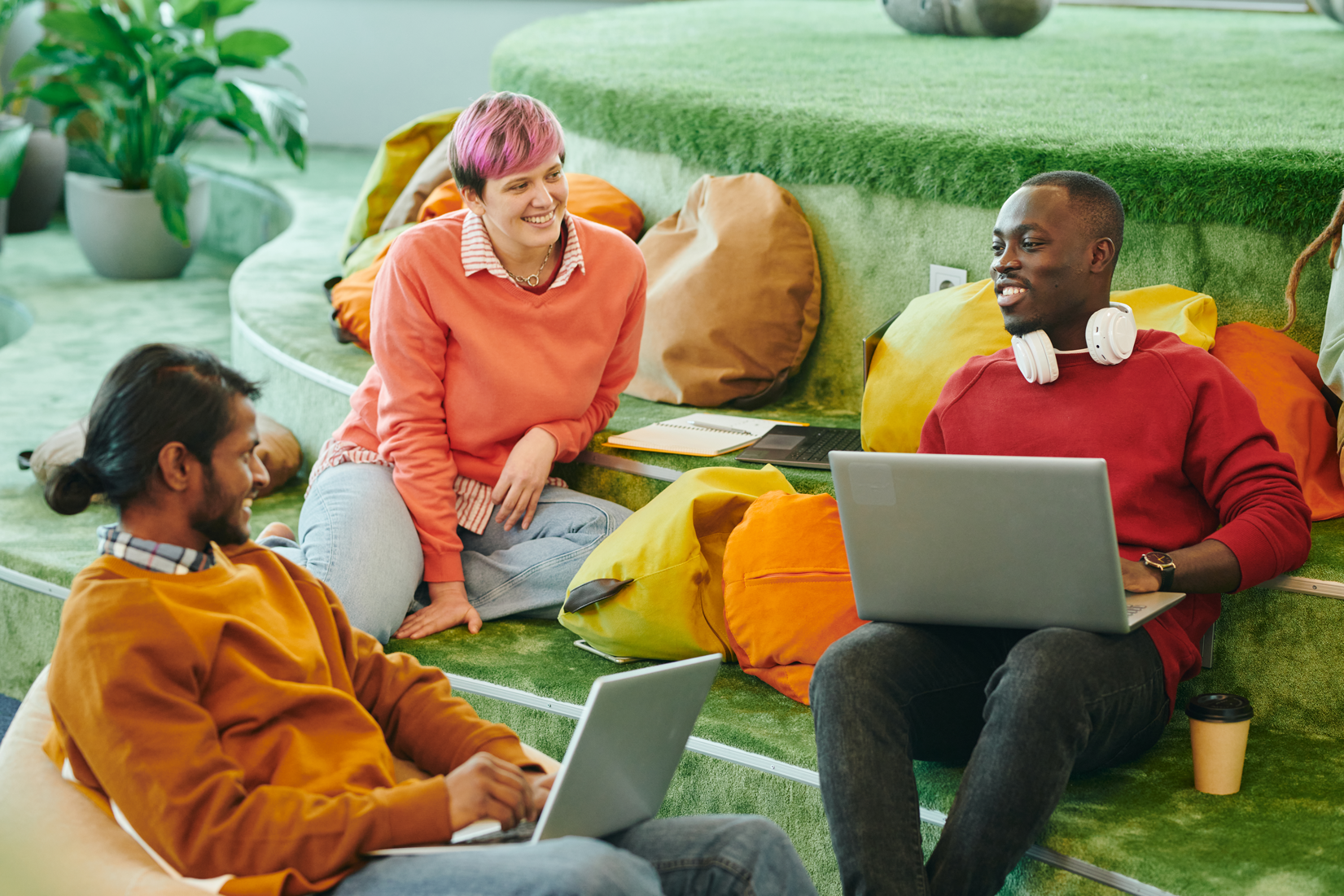 Four people sitting on green carpeted stairs in a casual setting, engaging in conversation with laptops and notebooks, with vibrant cushions and potted plants.