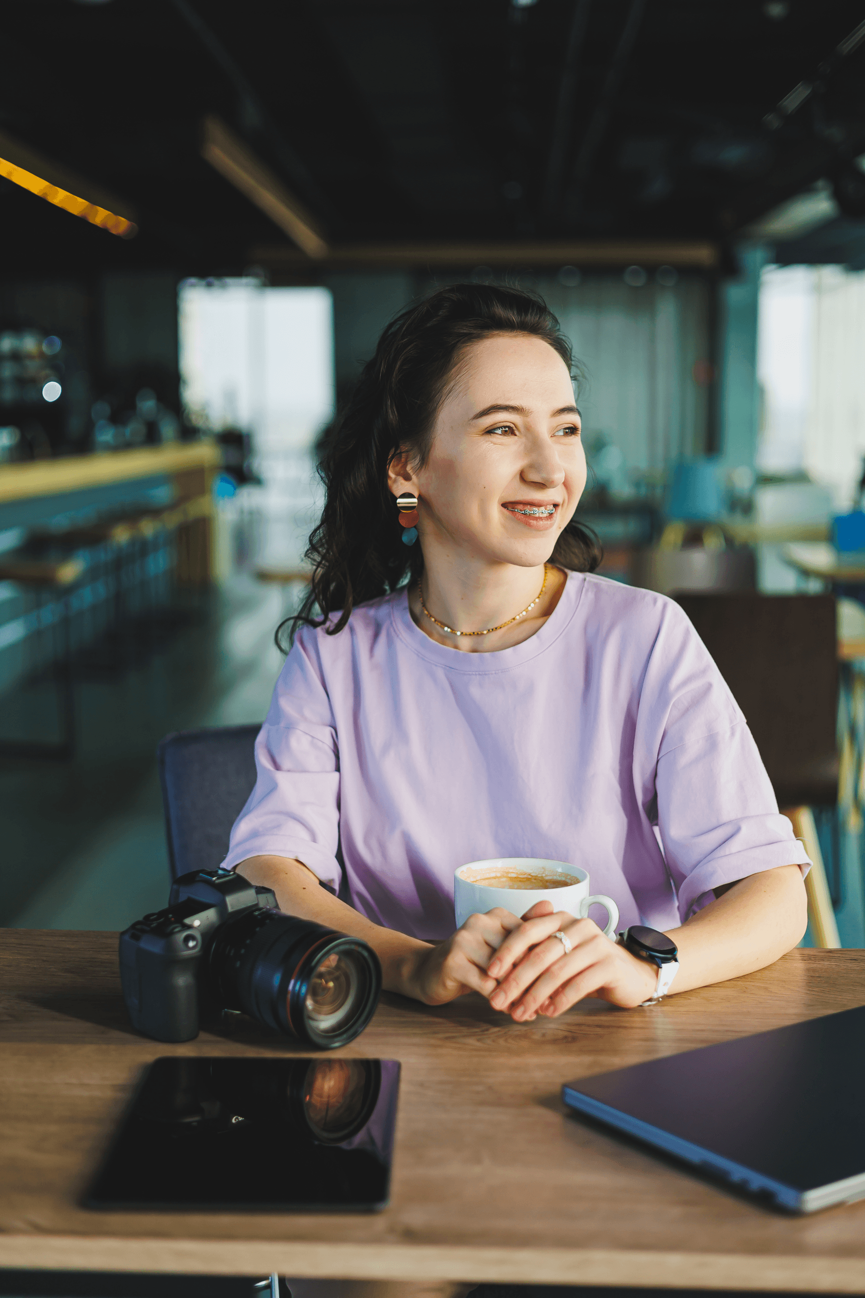 A young woman with dark curly hair and braces smiling while sitting at a wooden table with a camera, a tablet, a laptop, a cup of coffee, and a smartwatch in a modern cafe with large windows.