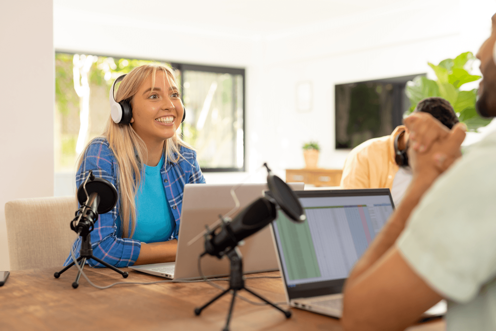 A group of people in a podcast or radio recording session, with a smiling woman wearing headphones in focus, microphones, laptops, and a bright, modern room with large windows and green plants.