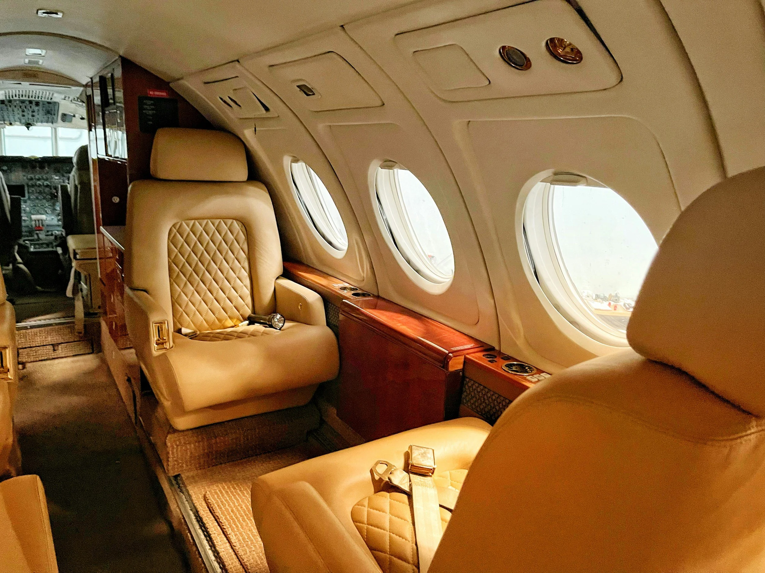Inside a private jet with beige leather seats, small windows, and wooden accents. The cockpit is visible at the front of the aircraft.
