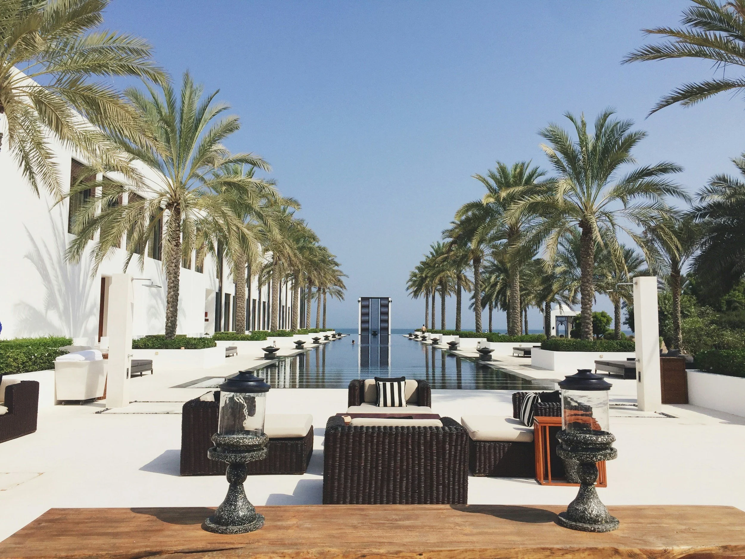 An outdoor seating area with black wicker chairs and tables with marble tops, set around a reflecting pool, with palm trees lining a white building and a clear blue sky in the background.