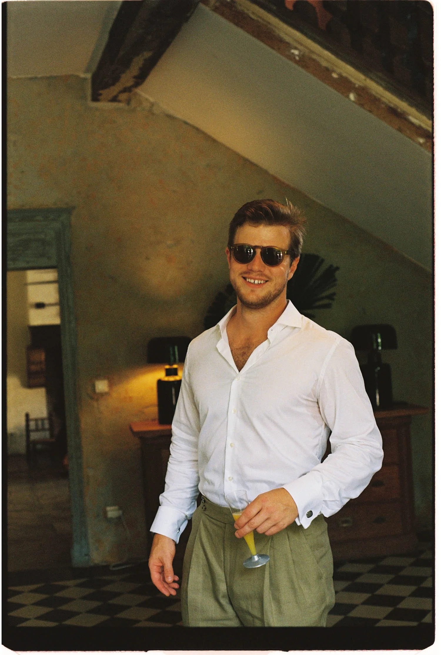 A young groom wearing sunglasses and a white shirt, smiling and holding a cocktail with a yellow drink and a lemon slice in a glass, at an indoor hall.
