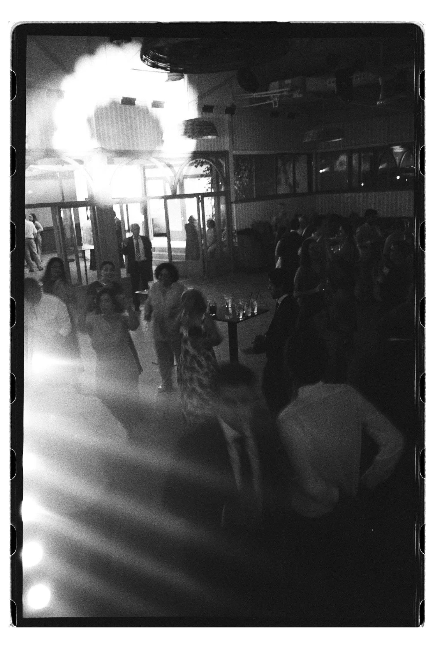 Analogue black-and-white photo of a crowded indoor wedding party with people mingling and dancing. Lights, movement and blur feeling give the photo an ethereal look.