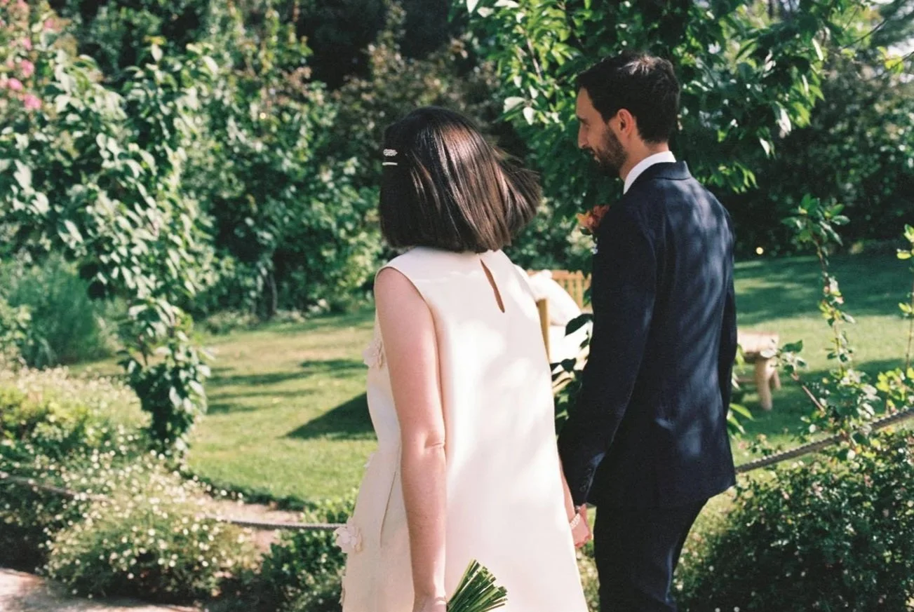 A bride and groom holding hands and smiling at each other outdoors in a garden setting during a wedding ceremony.