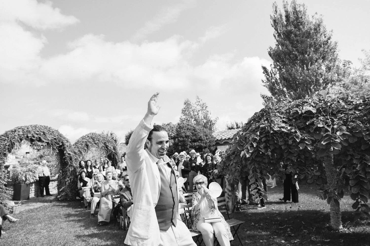 Groom in a suit and light-colored jacket, smiling and waving at an outdoor wedding ceremony, with a group of people seated and standing in the background, under a partly cloudy sky in a garden.