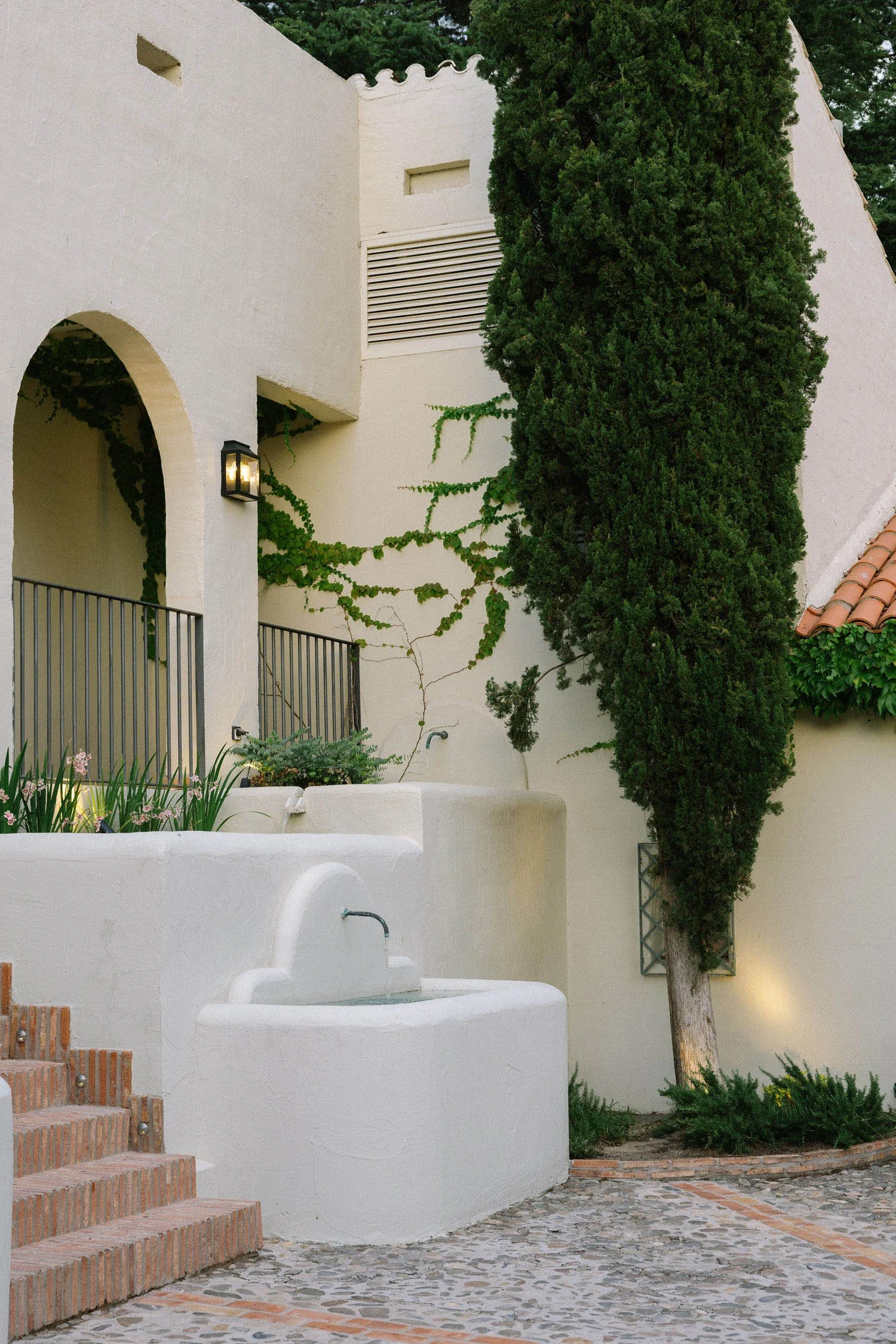 Exterior of a building with white stucco walls, a large bushy tree, stone pathway, and a brick staircase with metal railing. The building features an arched entryway, a wall-mounted lantern, and a small window. The scene suggests a Mediterranean or s