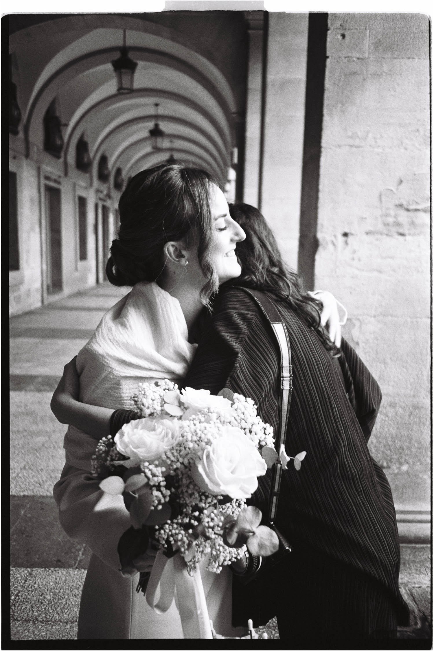 Two women embracing, one holding a bouquet of flowers, in an arched outdoor corridor, black and white photograph.