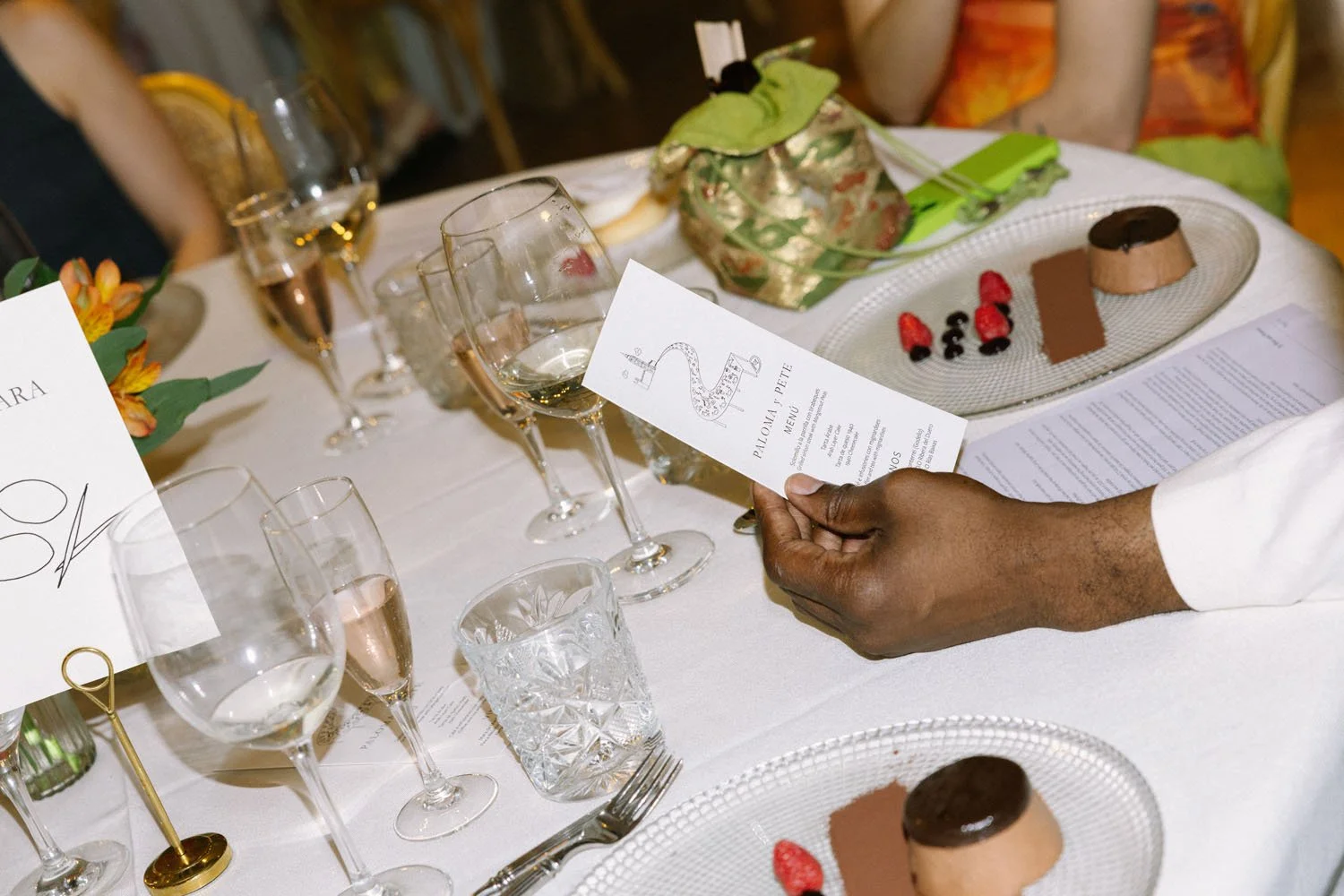 A person holding a menu at a formal dinner table with glasses of water and wine, floral centerpiece, and plates with desserts and berries.
