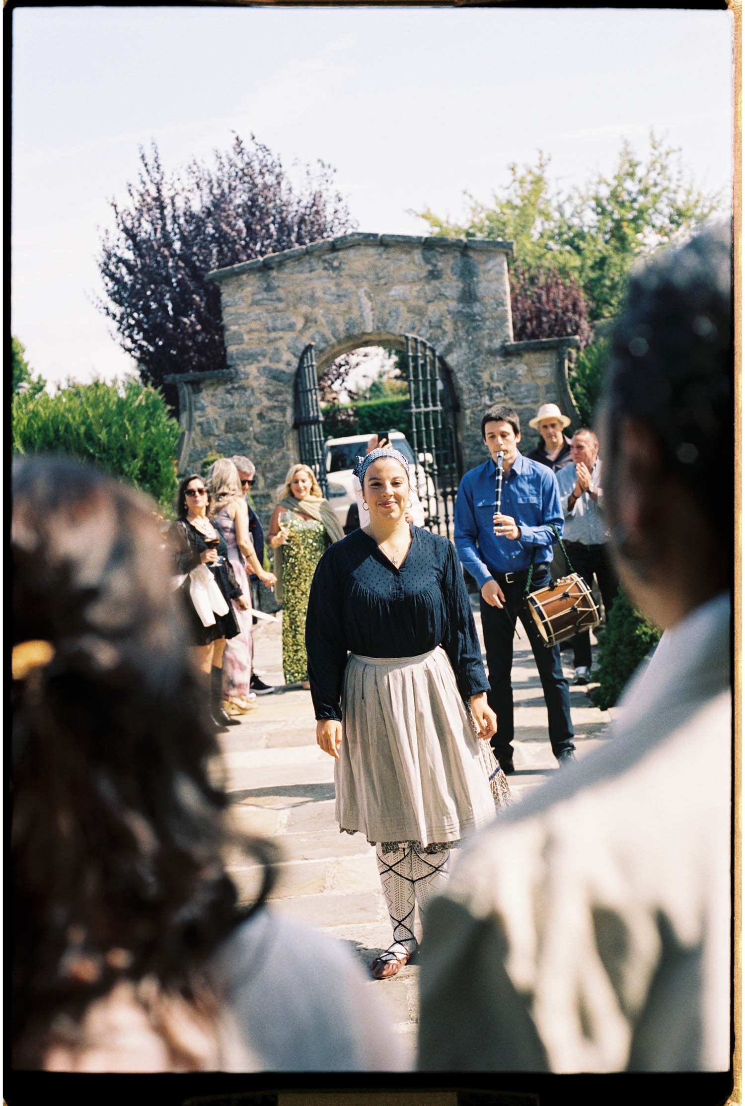 A woman in traditional Basque vintage clothing dancing through a wedding crowd for the just-married couple, with a stone gate and trees in the background.