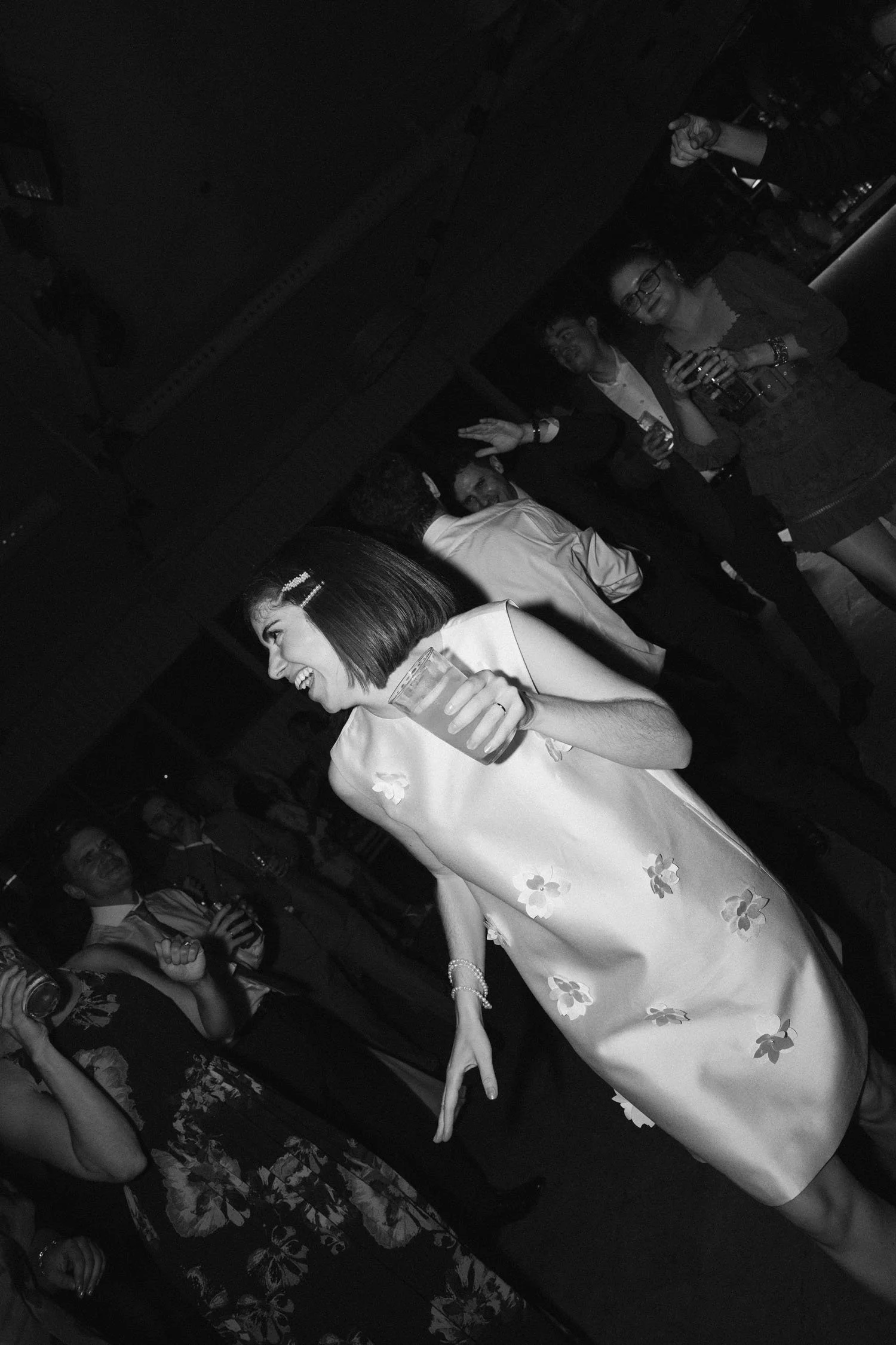 Bride celebrating her wedding party, she wears a floral dress in the foreground holding a drink and smiling, surrounded by guests and enjoying the event.