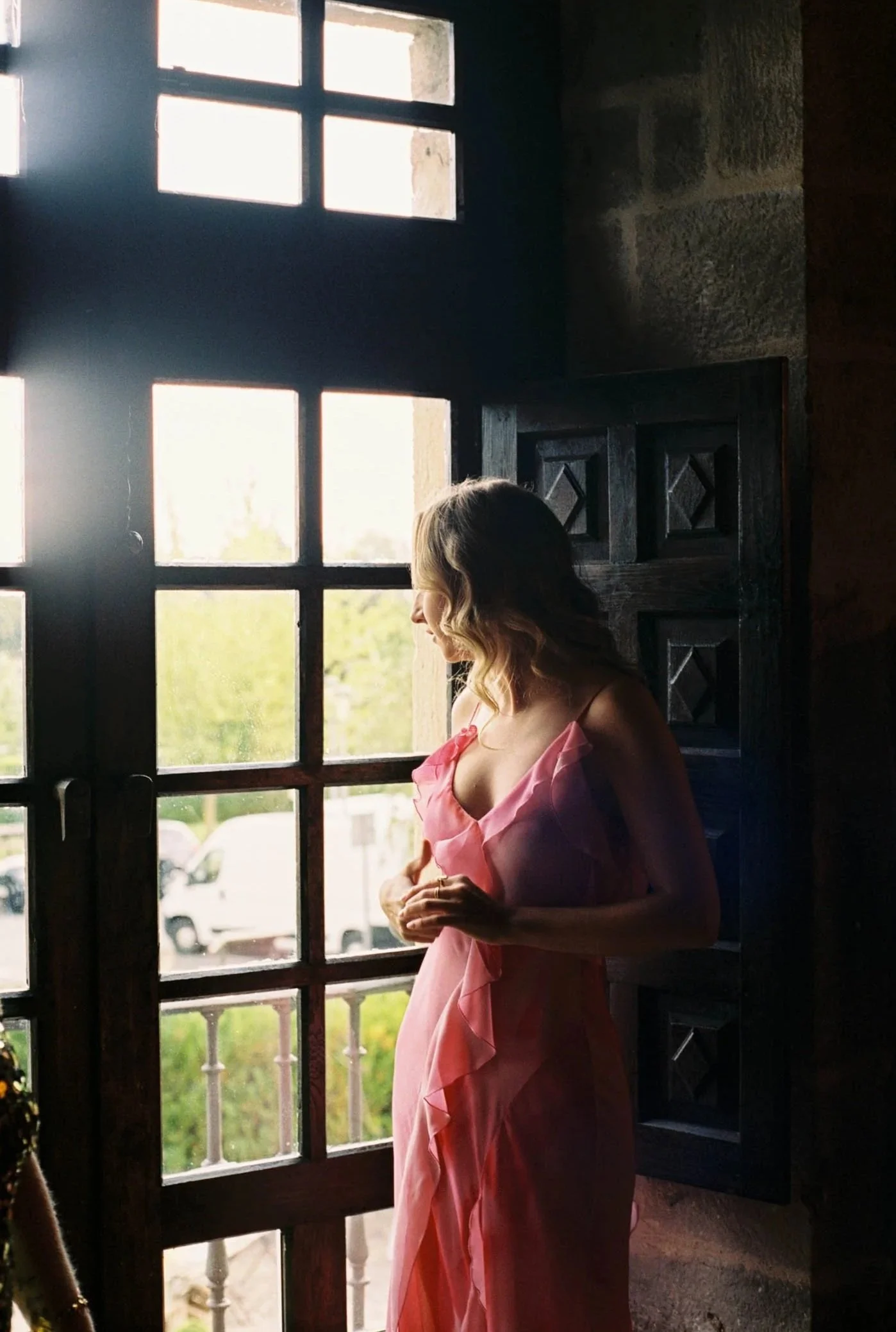 Analogue cinematic photo of a wedding guest in a pink dress standing by a window with sunlight streaming in, looking outside.