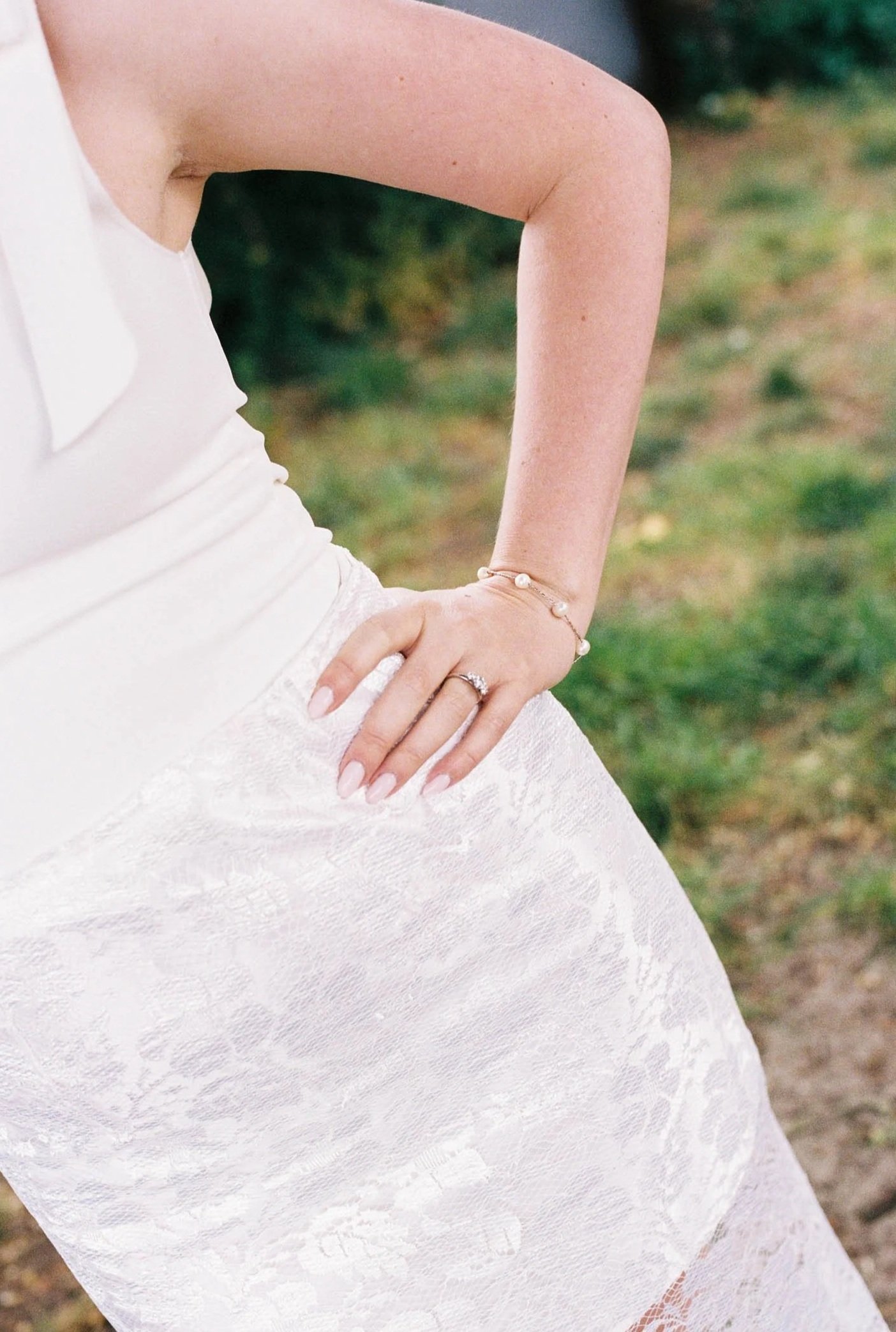 Close-up of a person wearing a white lace dress or skirt, with one hand resting on their hip, outdoors with grass and trees in the background.