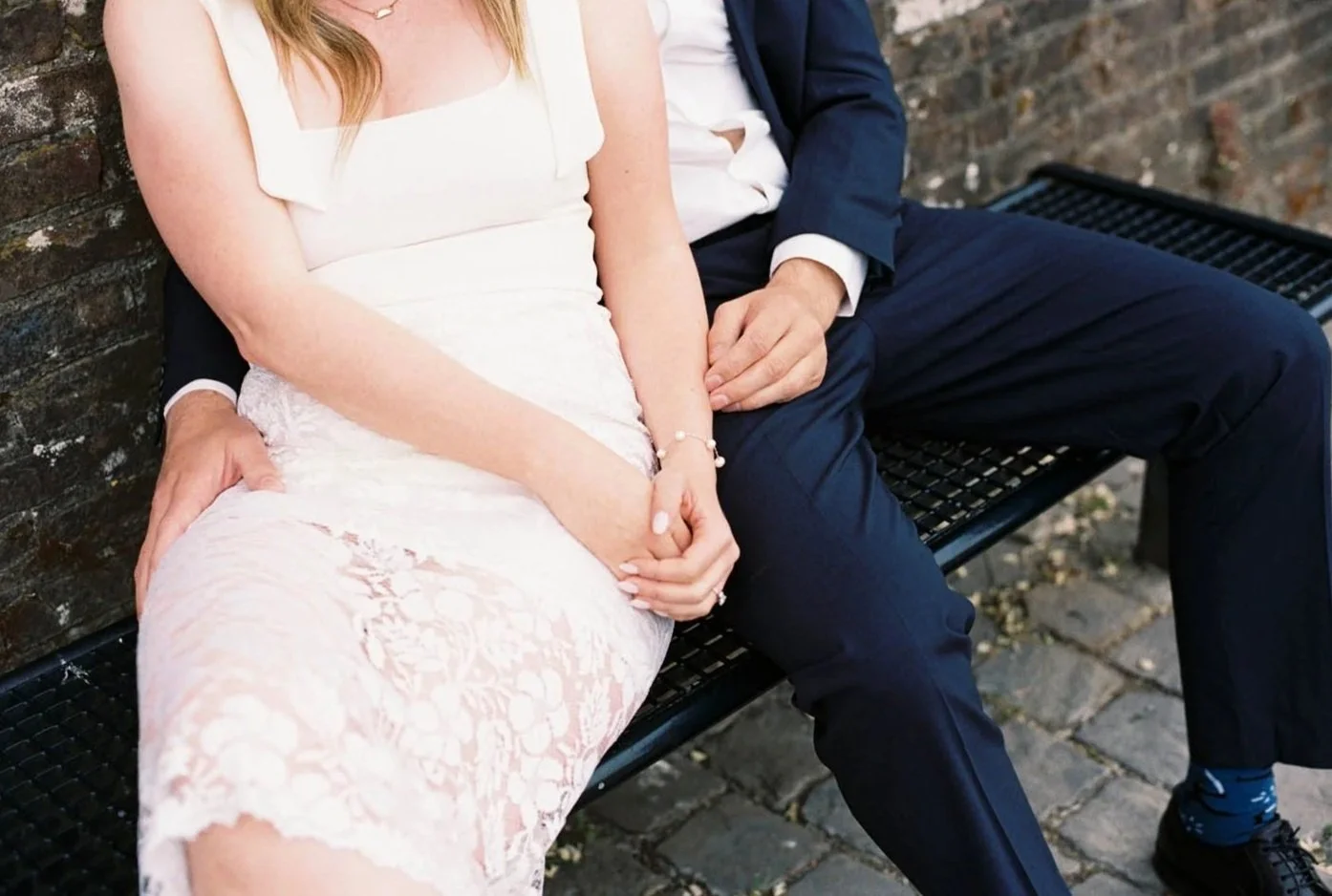 A couple sitting on a bench, with the woman in a white dress and the man in a navy suit, holding hands. The woman is wearing a lace dress and jewelry, and they are sitting on a black metal bench against a brick wall.