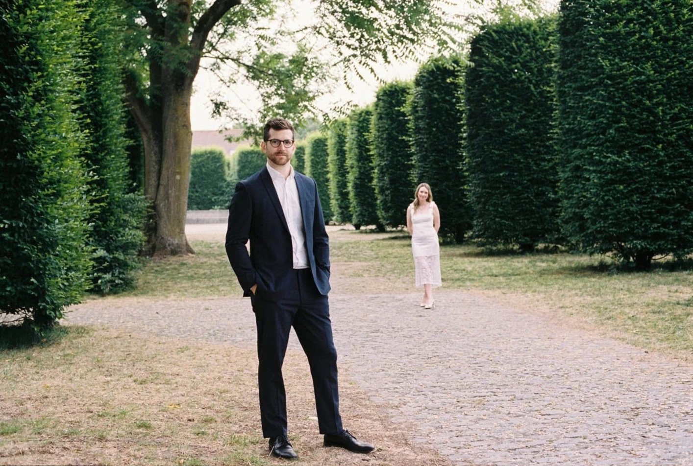 A man in a dark suit stands with his hands in his pockets in the foreground, a woman in a white dress stands further back on a gravel path in a park-like setting with tall, dense shrubbery and trees.