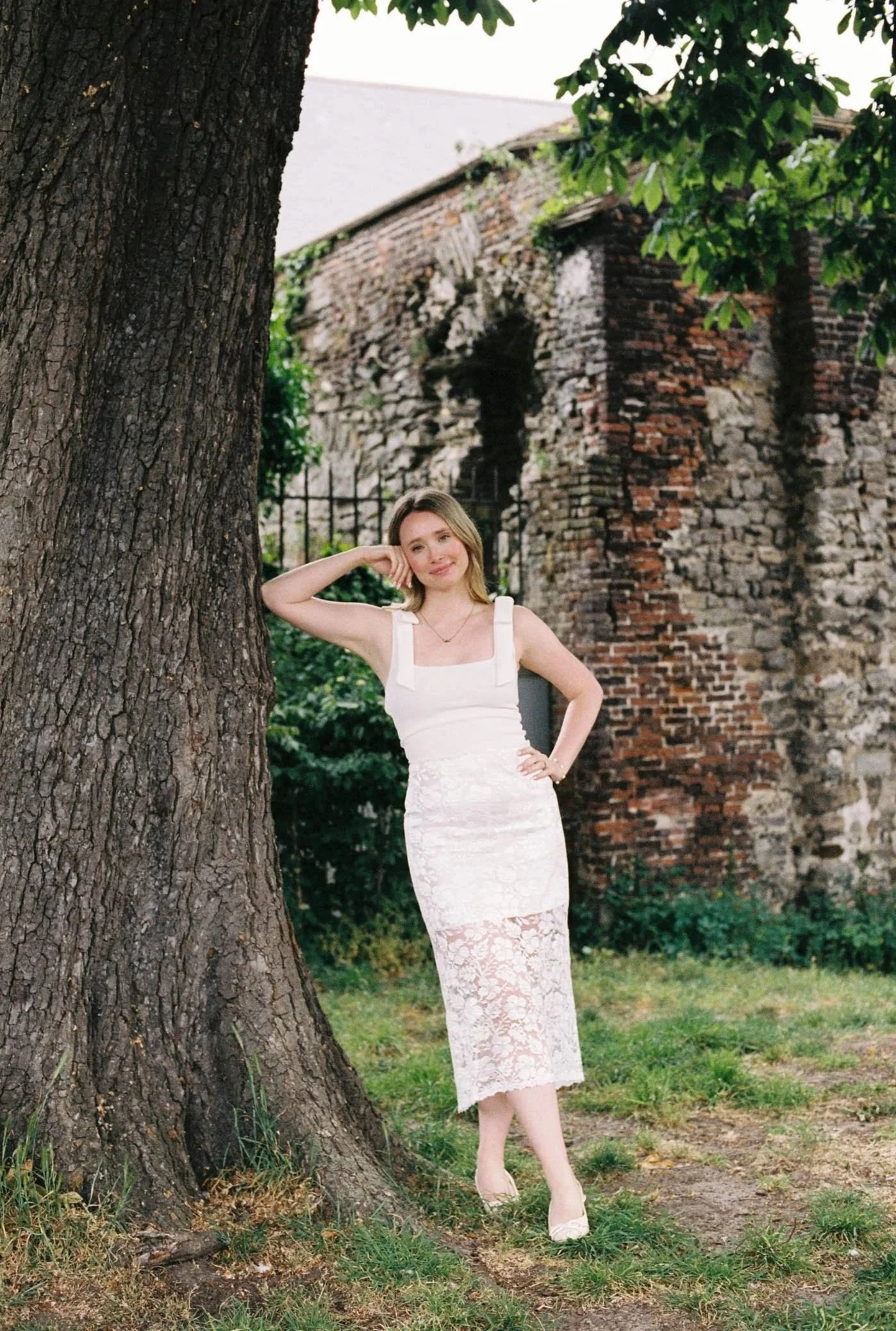 A woman in a white lace dress standing beside a large tree with an old brick building in the background.