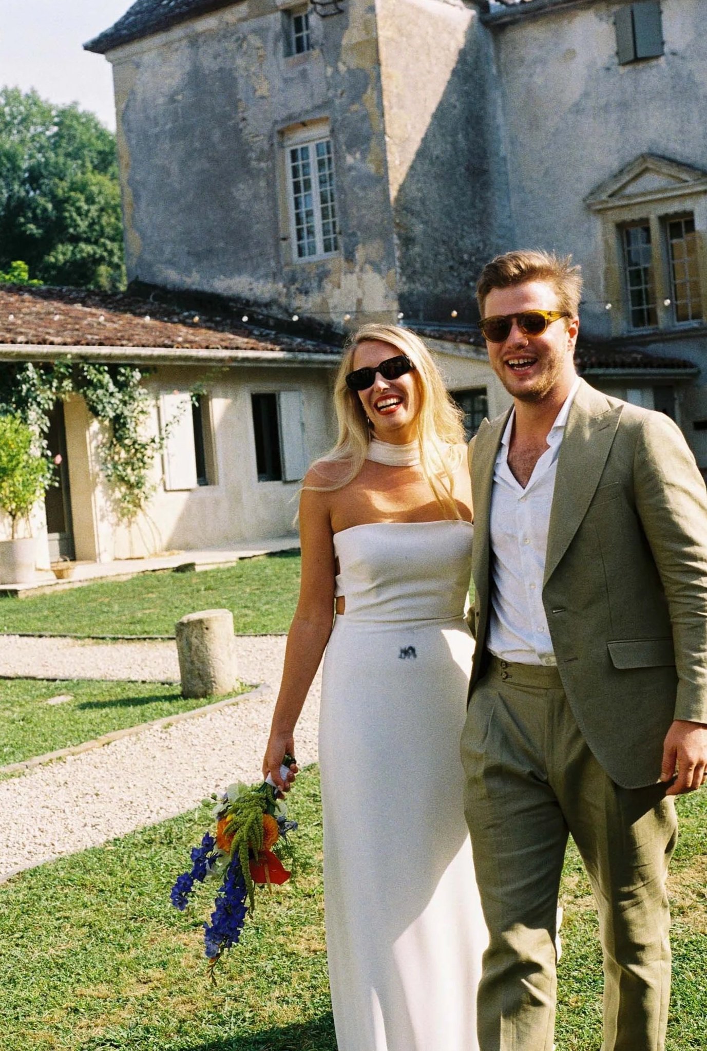 A smiling couple dressed in wedding attire standing outdoors in front of an old stone building, with a woman holding a bouquet of colorful flowers.