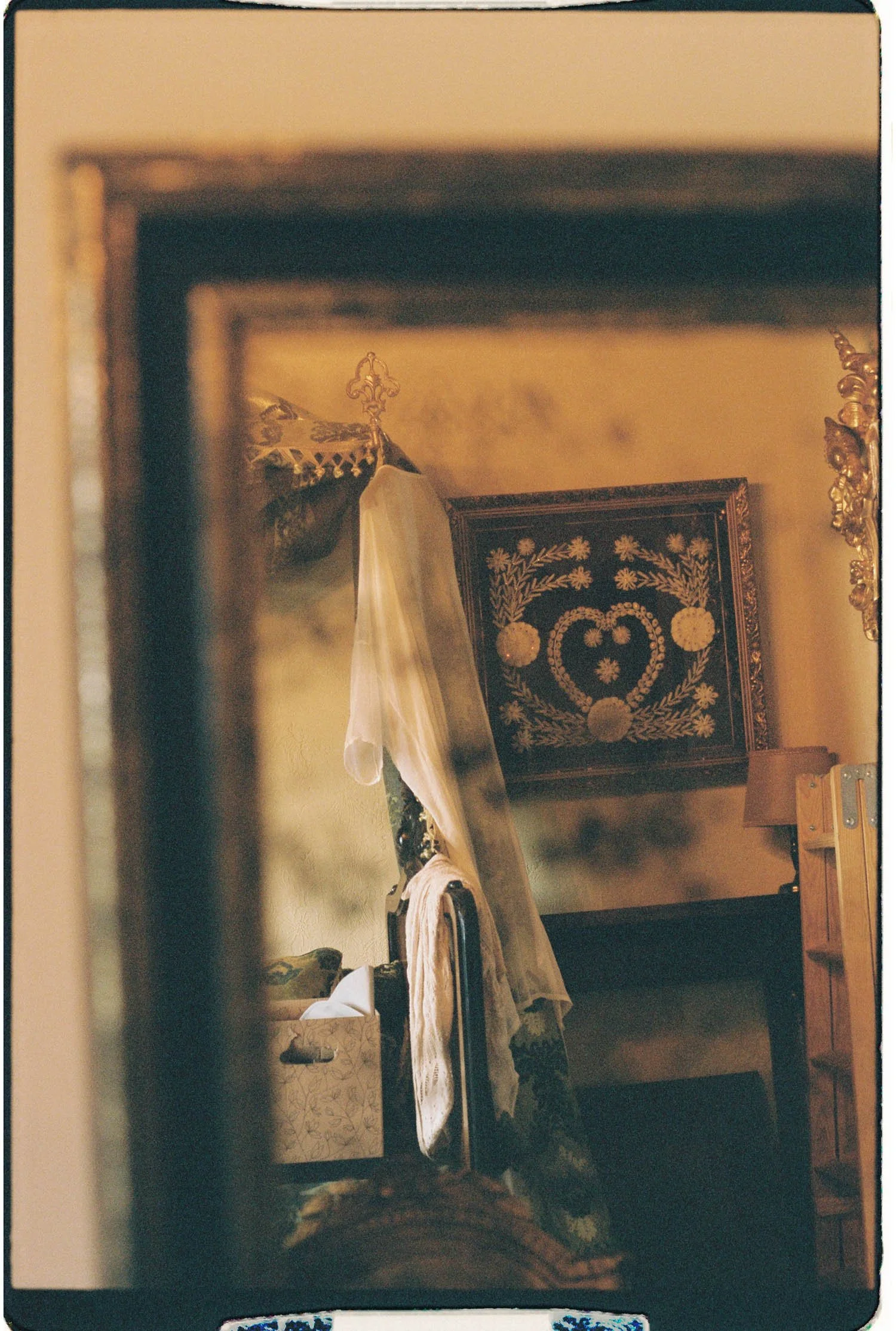 Analogue image of a bedroom with a bed covered in white sheets and a decorative headboard. Bride's dresser with a patterned box on top, a wall hanging with floral patterns, and various items on the floor including clothes and shoes.