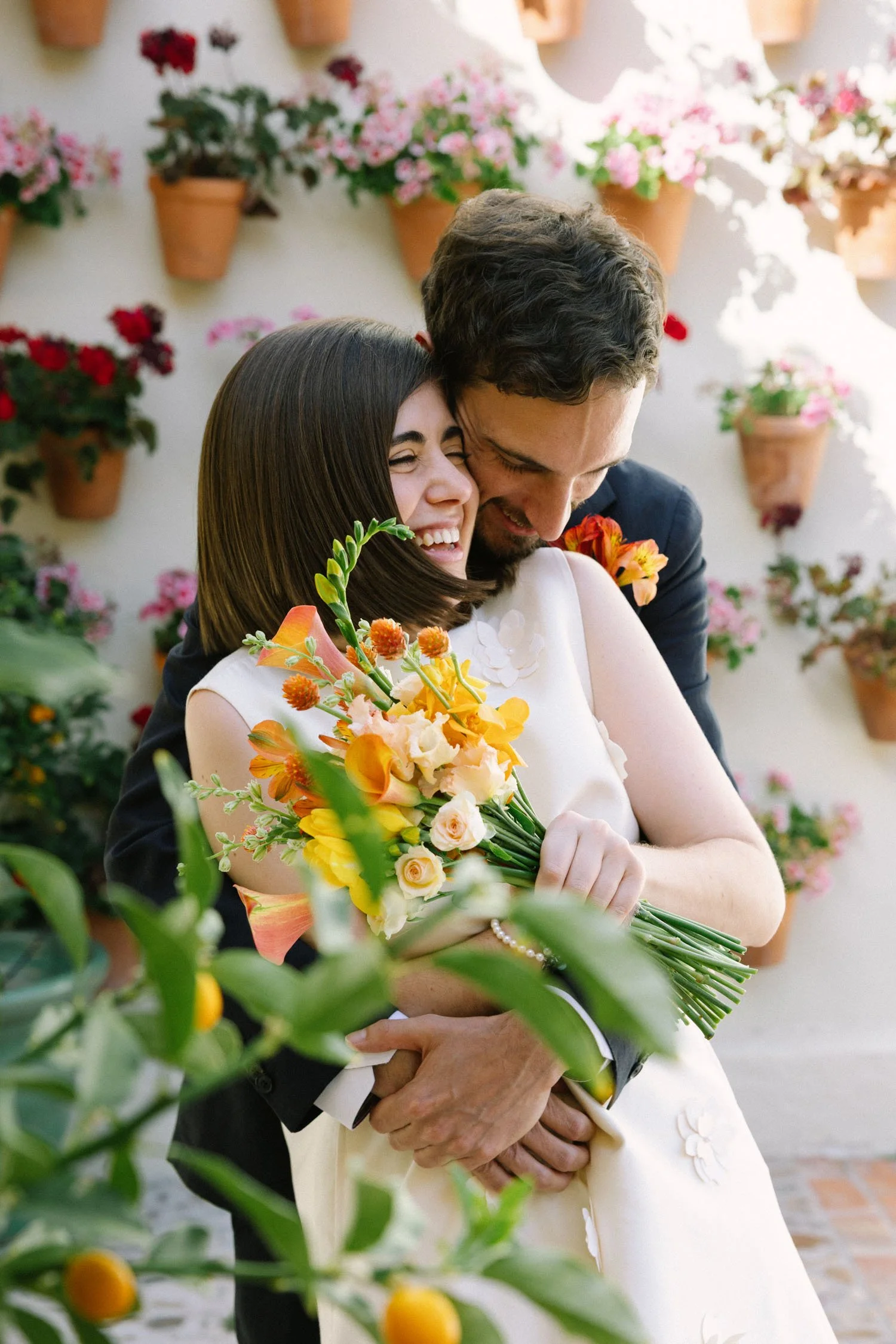 A bride and groom embrace in a garden with terracotta flower pots filled with pink and red flowers on a white wall behind them. The bride holds a bouquet of orange, yellow, and pink flowers, and both appear joyful and happy.