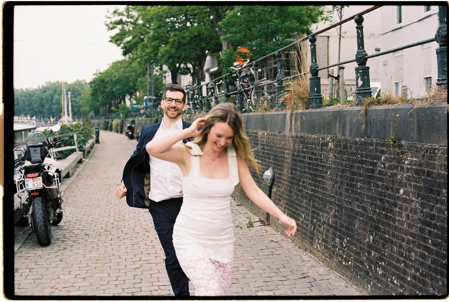 Analogue candid photo of a couple in wedding clothes walking along a cobblestone sidewalk by the water, with parked bicycles and trees in the background.