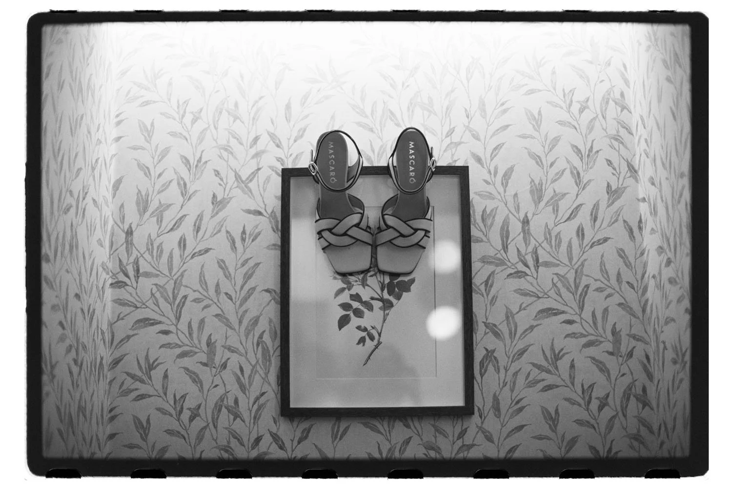 Pair of high-heeled sandals with woven straps displayed on a framed floral artwork mounted on a wall with leafy patterned wallpaper.