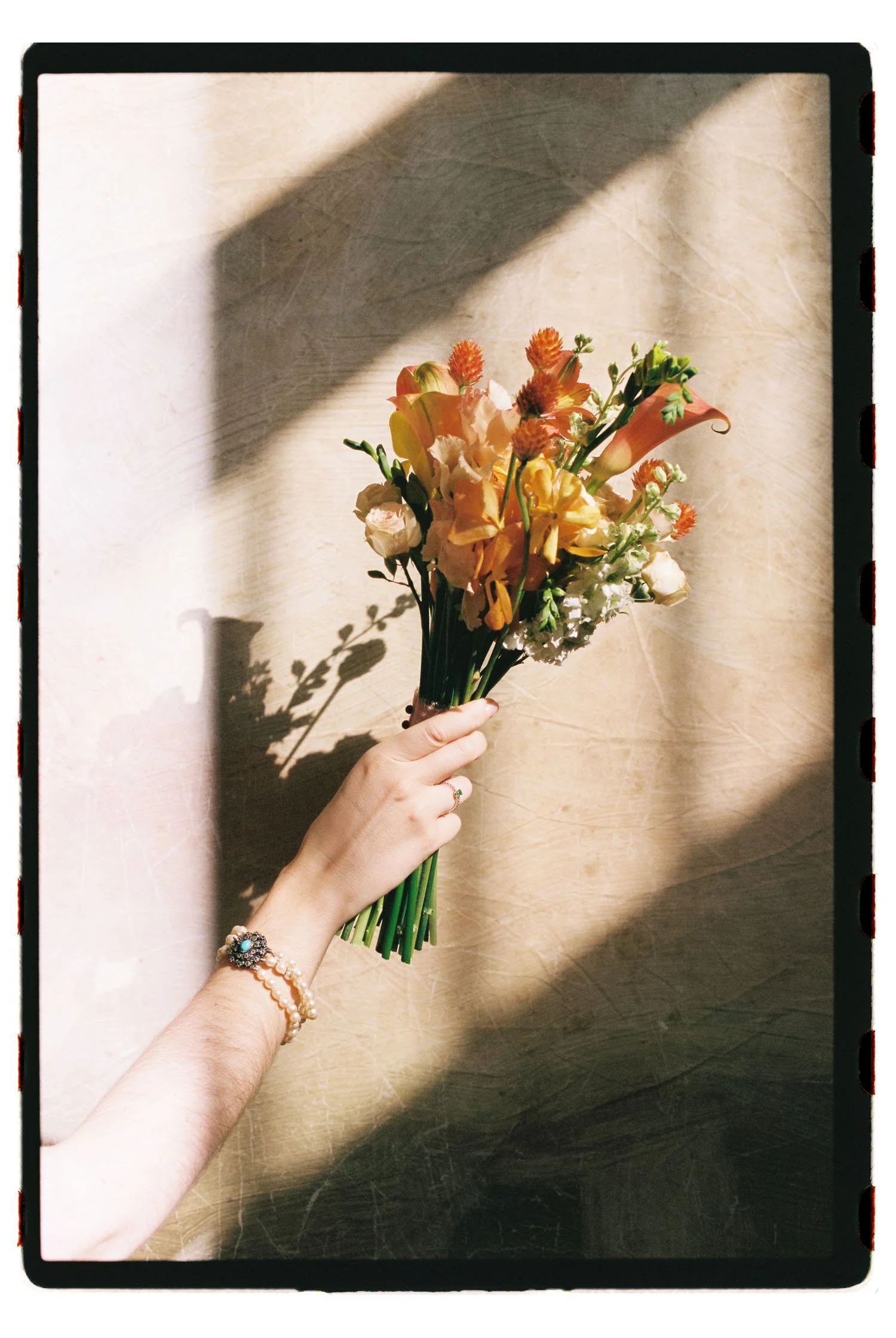 Analogue photo of a bride's hand holding a bouquet of various peach, white, and orange flowers with shadow cast on beige background.