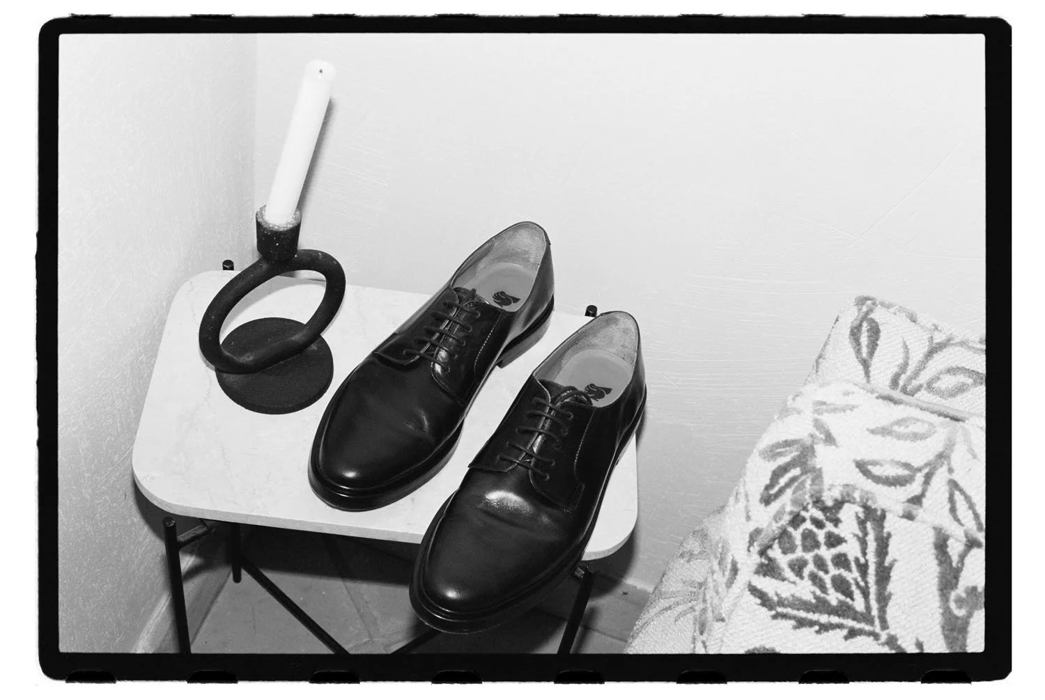 A pair of polished black dress shoes placed on a small white table, with a black kettlebell and a white candle in a holder nearby. A patterned fabric is partially visible in the bottom right corner.