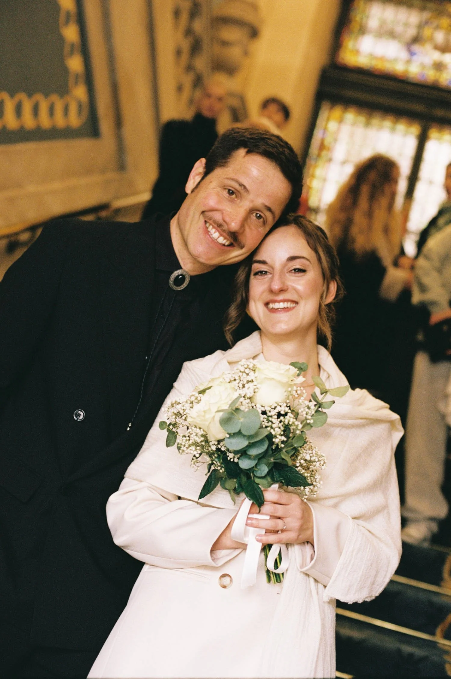 A man and woman smiling together at what appears to be a celebration or special event inside a decorated venue, with other guests in the background. The woman is holding a bouquet of white flowers and wearing a cream-colored dress, while the man is d
