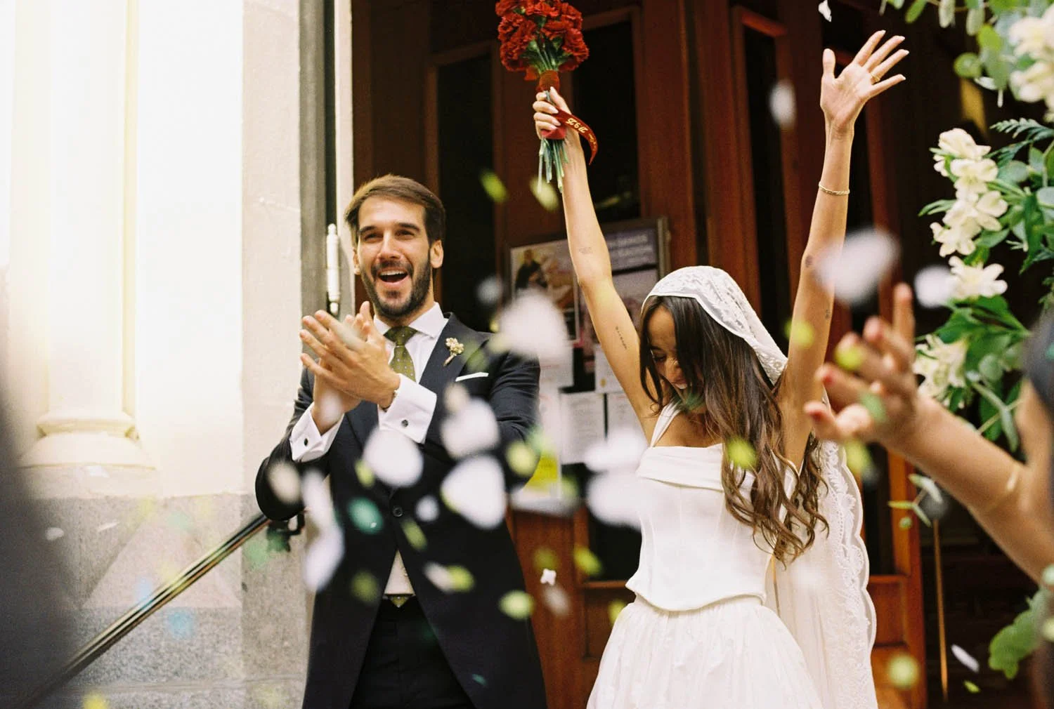 A joyful bride in a white dress and veil with her arms raised holding a bouquet, and a smiling groom in a suit clapping, celebrating inside a building with confetti falling around them.