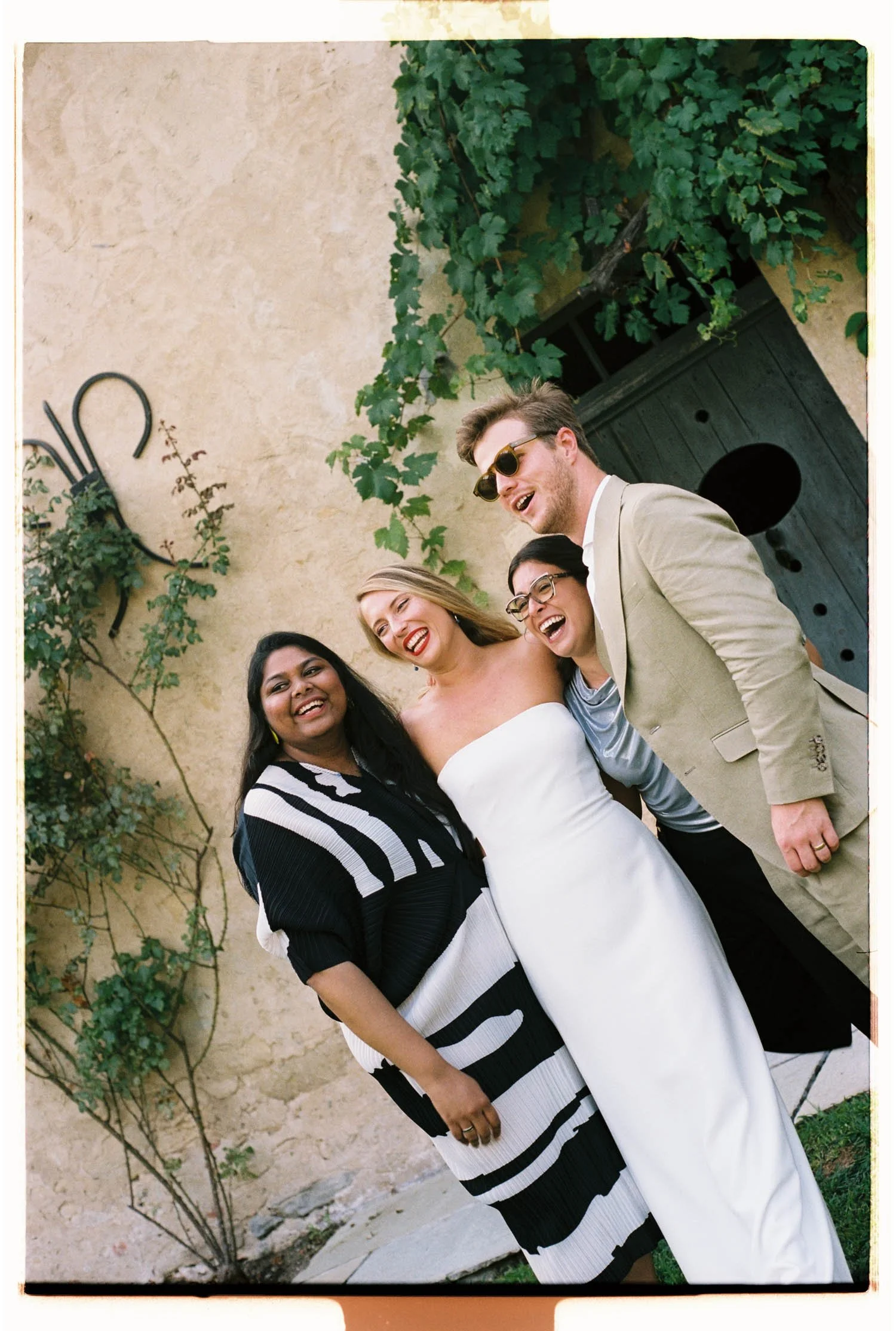 Group of diverse women and a man smiling and laughing outdoors in a wedding cocktail by a beige wall with green vines in a French chateau.