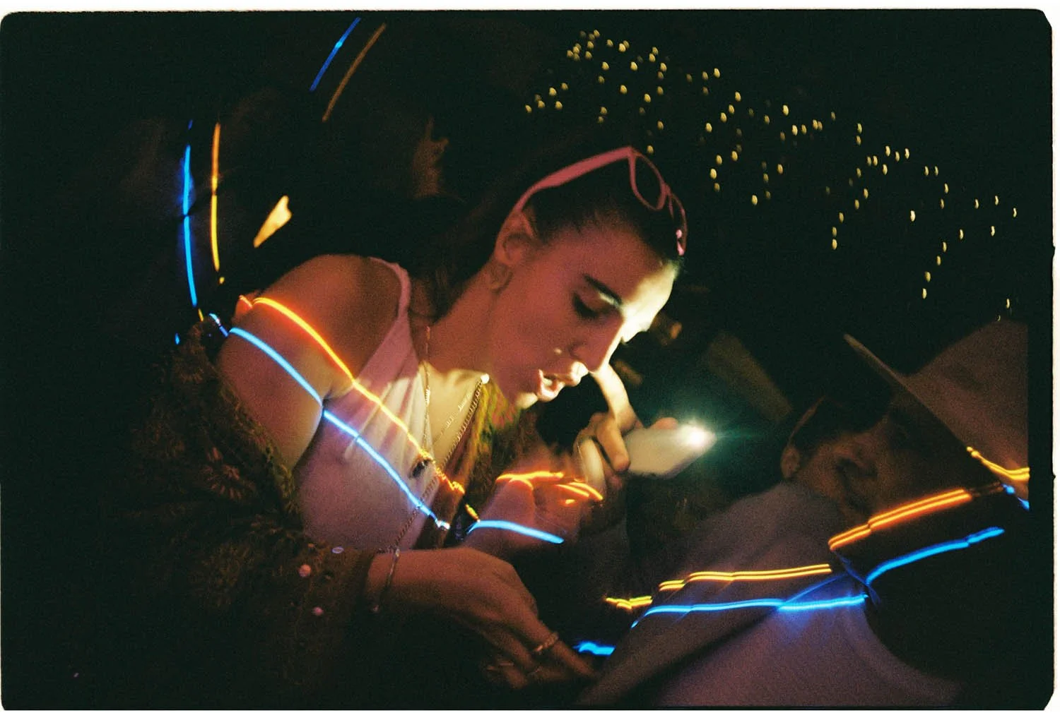 Analogue photo of a woman with glasses on her head looking at her phone with colourful light trails around her in a dark setting. In a wedding party at night.