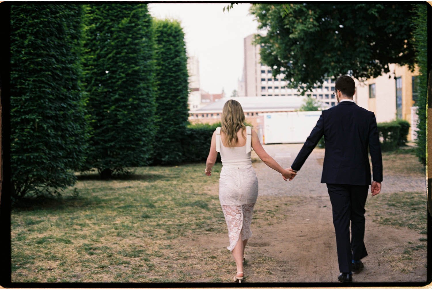 A couple walking hand in hand in an outdoor park area, with the woman wearing a white dress and the man wearing a suit.
