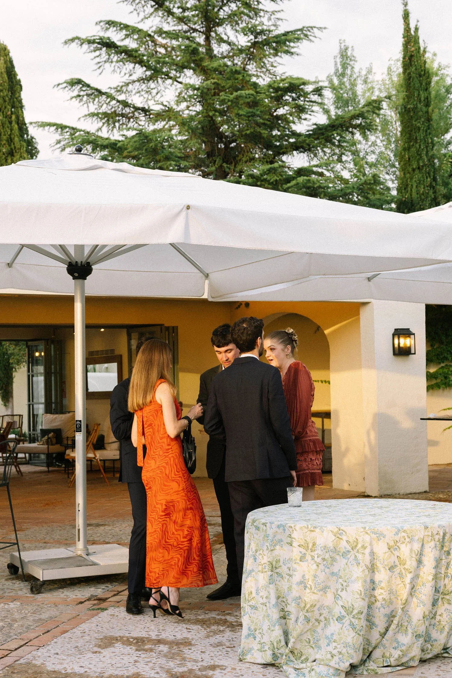 Group of wedding guests standing and talking outdoors under a white patio umbrella with trees and a building in the background.